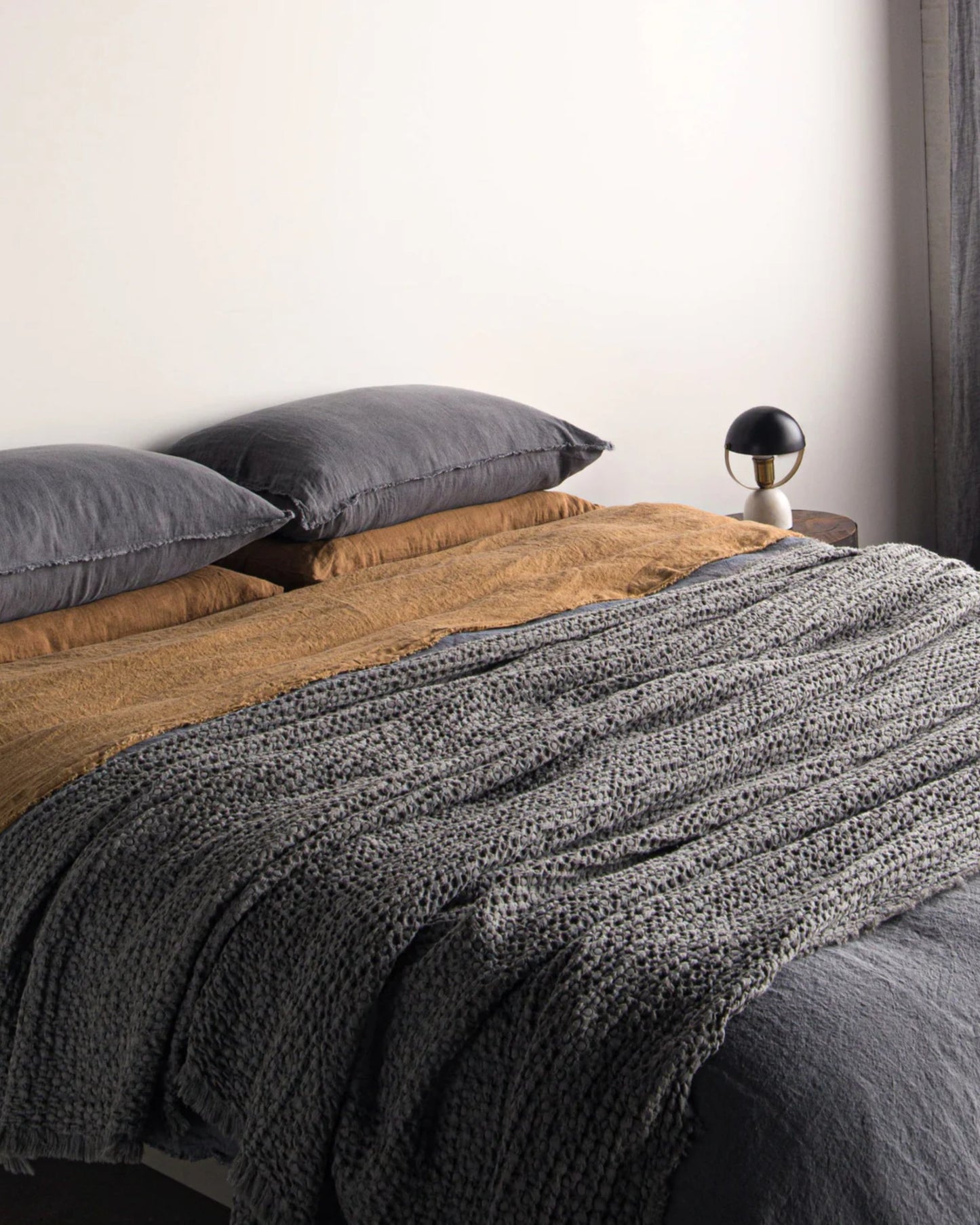 A neatly made bed with brown sheets and dark gray pillows showcases the HALE FLOCCA THROW from Hale Mercantile Co, adding a touch of elegance. A small black and white lamp sits on a nightstand next to the bed, set against a crisp white wall.