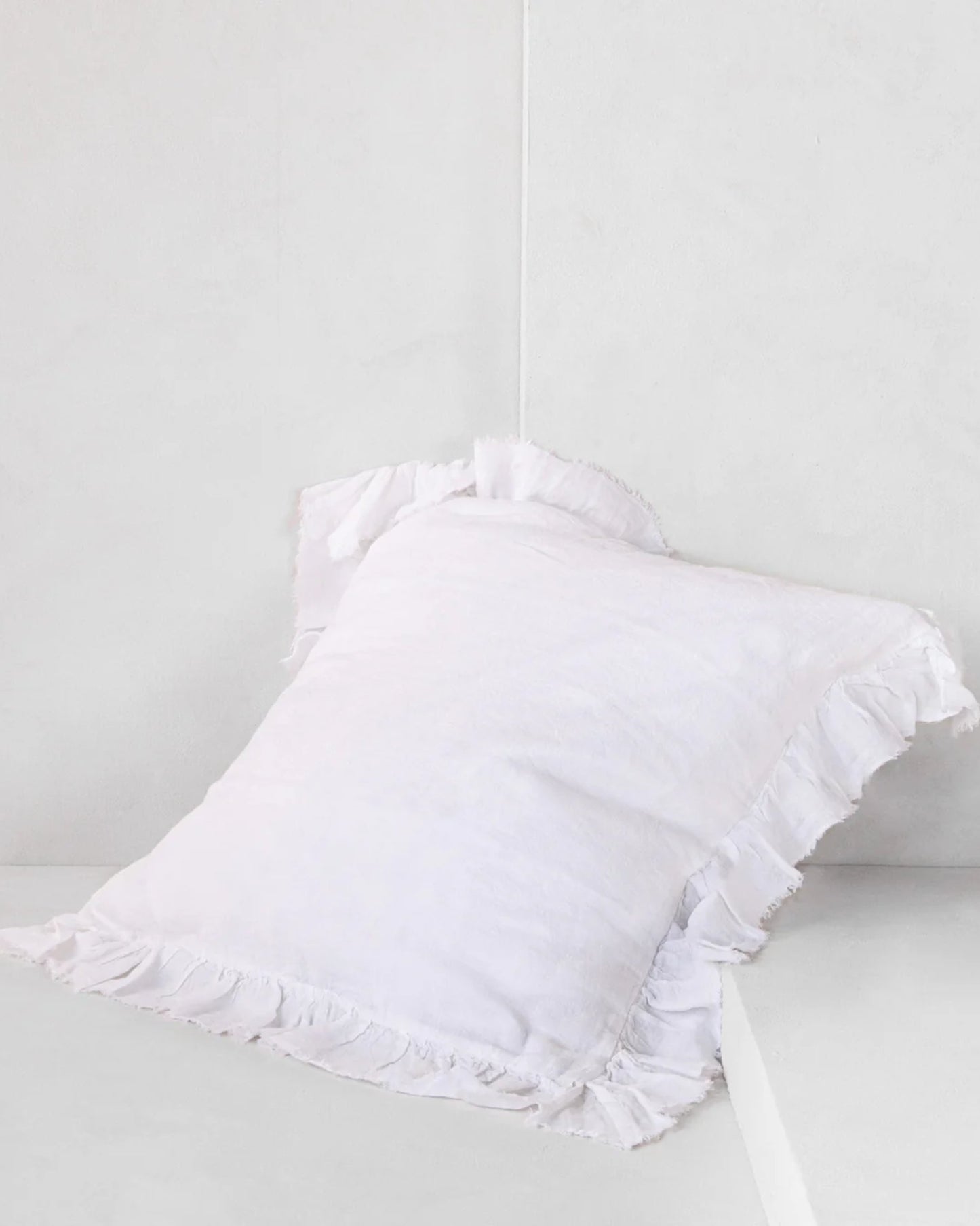 The HALE KRISTINE EURO PILLOWCASE by Hale Mercantile Co, featuring a classic white design with ruffled edges that evoke a vintage-inspired bedroom aesthetic, is positioned against a light gray wall on a white surface.