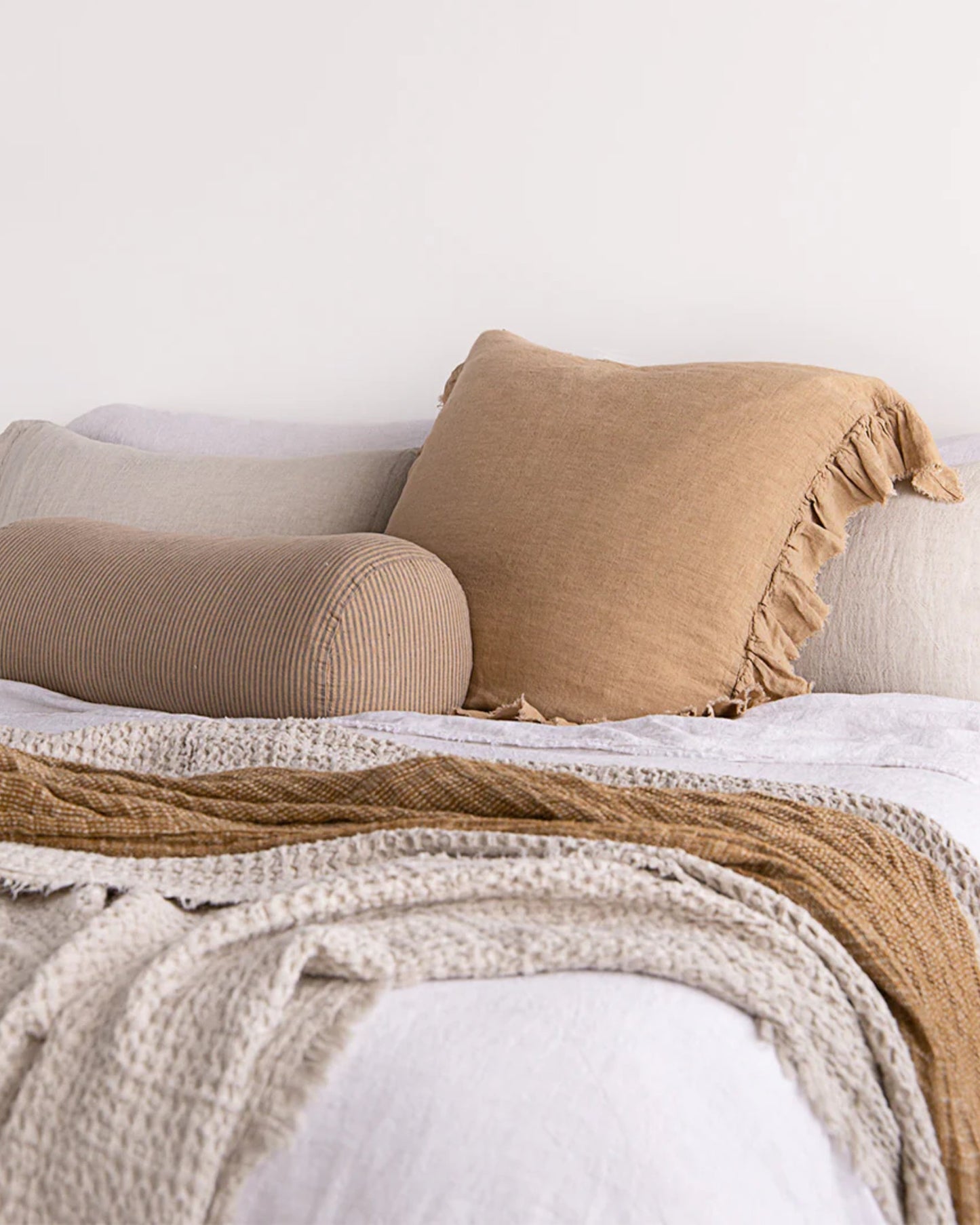 A neatly made bed features the HALE KRISTINE EURO PILLOWCASE by Hale Mercantile Co, showcasing a beige square pillow with fringe, complemented by a cylindrical striped pillow and a layered knit blanket in shades of white and tan. This vintage-inspired bedroom decor creates a cozy and inviting atmosphere.