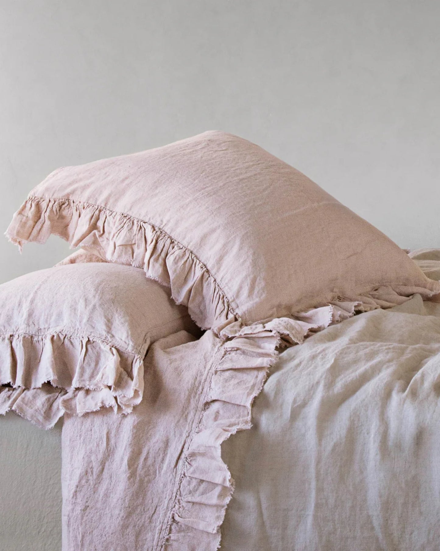 A set of two HALE KRISTINE EURO PILLOWCASES by Hale Mercantile Co in beige with ruffles is elegantly arranged on a coordinating duvet, enhancing the room with a vintage-inspired bedroom decor. The soft, textured linen fabric contributes to a cozy ambiance against the plain off-white wall.