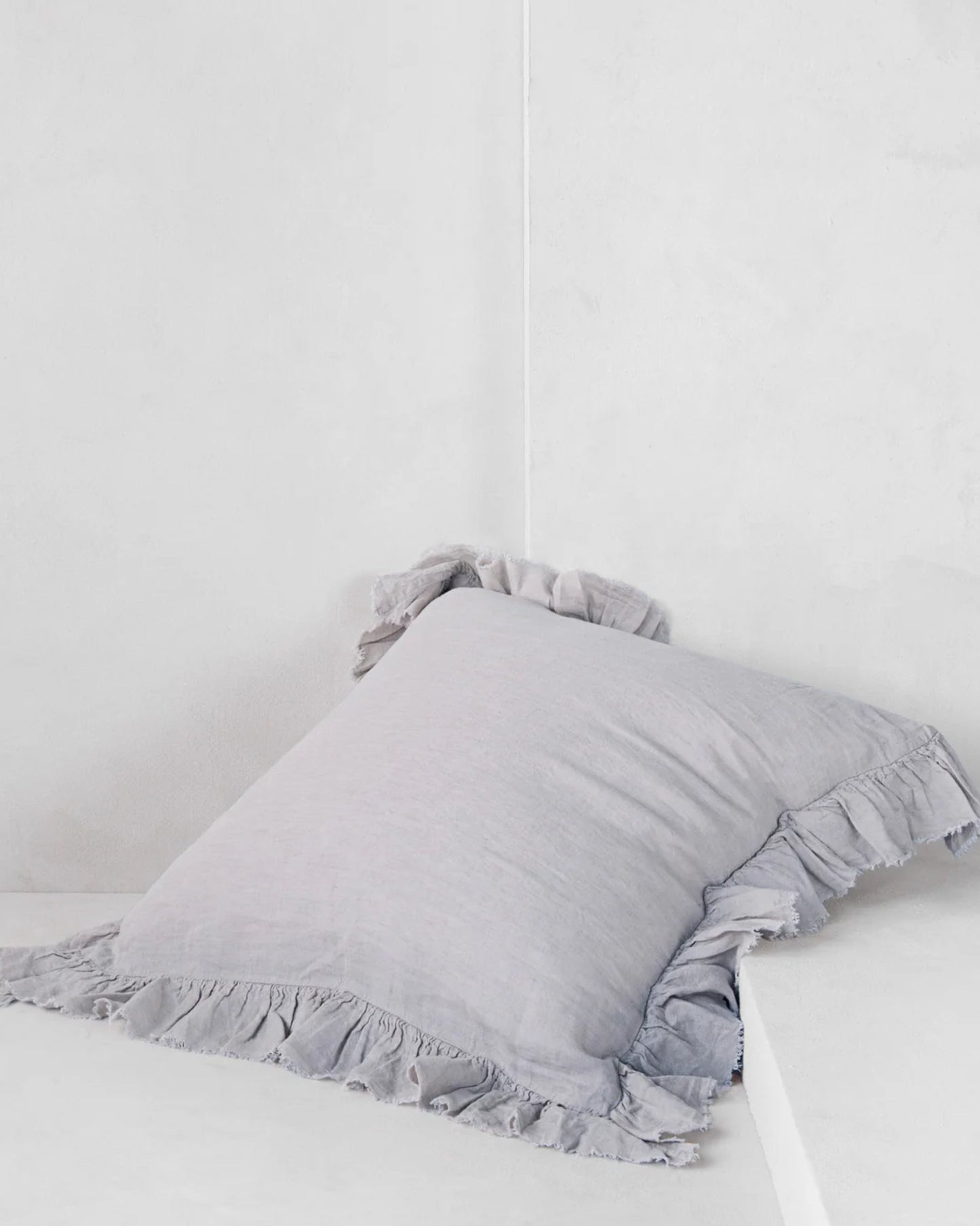 A soft, light gray Hale Kristine Euro Pillowcase with ruffled edges, evoking vintage-inspired bedroom decor from Hale Mercantile Co, rests against a white corner wall.