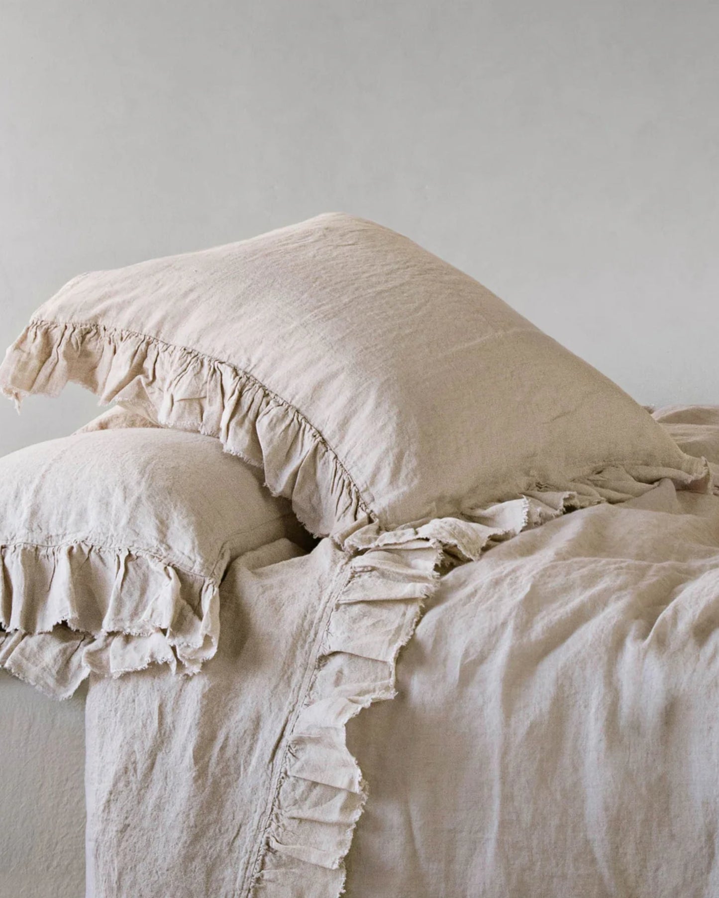 The HALE KRISTINE EURO PILLOWCASE by Hale Mercantile Co, featuring a beige color and ruffled edges, rests on a neatly arranged bed paired with matching vintage-inspired bedding. The easy-care linen fabric offers a natural, textured look that adds a cozy and rustic feel to the setting.