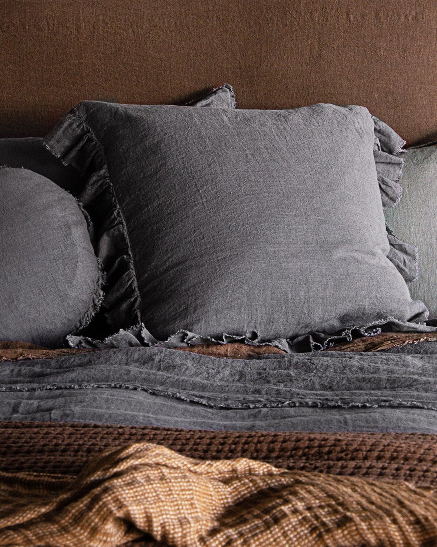 A cozy bedroom setting featuring a bed with the HALE KRISTINE EURO PILLOWCASE from Hale Mercantile Co., adorned in ruffled gray European linen and complemented by layered textured blankets in shades of brown and gray, creating a warm and inviting atmosphere.