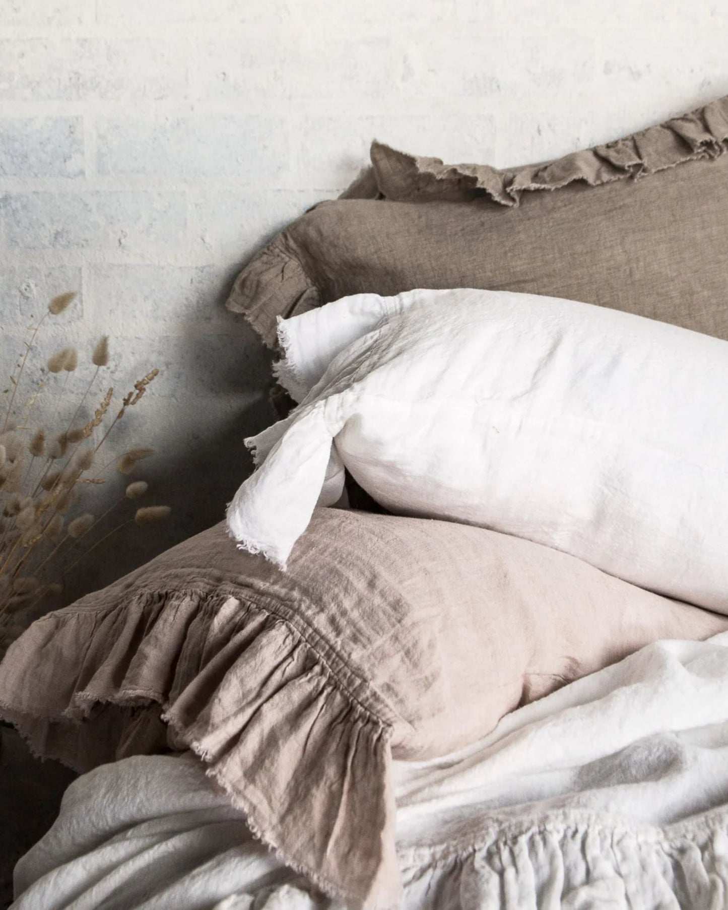 A cozy bed setup highlights a stack of HALE KRISTINE EURO PILLOWCASES by Hale Mercantile Co, featuring earthy tones with ruffled edges, perfectly paired with an easy-care linen bedding set. The light-colored, textured wall and dried flowers enhance the vintage-inspired bedroom decor's rustic ambiance.