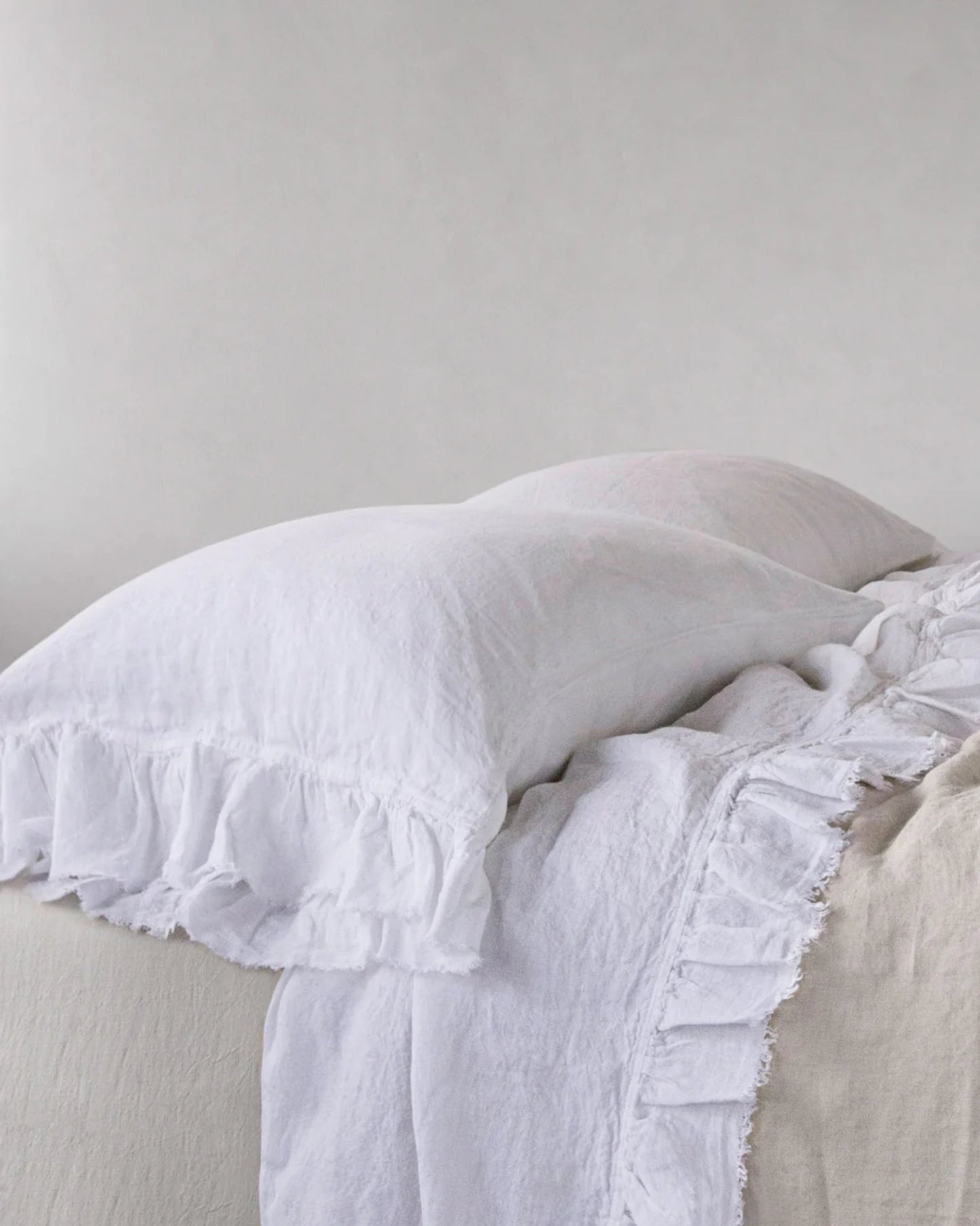 A neatly made bed showcases two white pillows and a white ruffled bedspread over a beige blanket, offering a vintage charm. The bedding's soft, textured look enhances the cozy and inviting atmosphere, with HALE KRISTINE STANDARD PILLOWCASES by Hale Mercantile Co adding to the rustic romance of the scene.