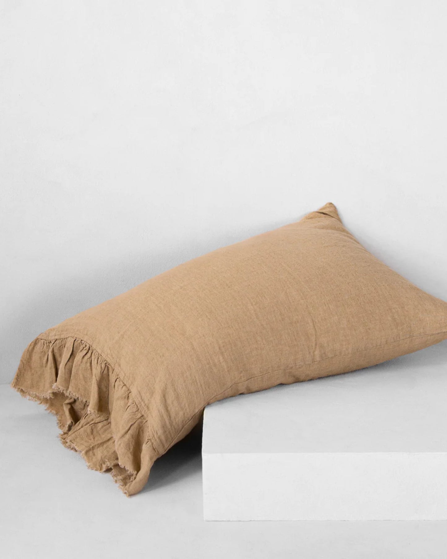 The HALE KRISTINE STANDARD PILLOWCASE by Hale Mercantile Co, in a beige rectangular design featuring ruffled edges on one side, exudes rustic romance as it lies against a plain white background, partially atop a white block.