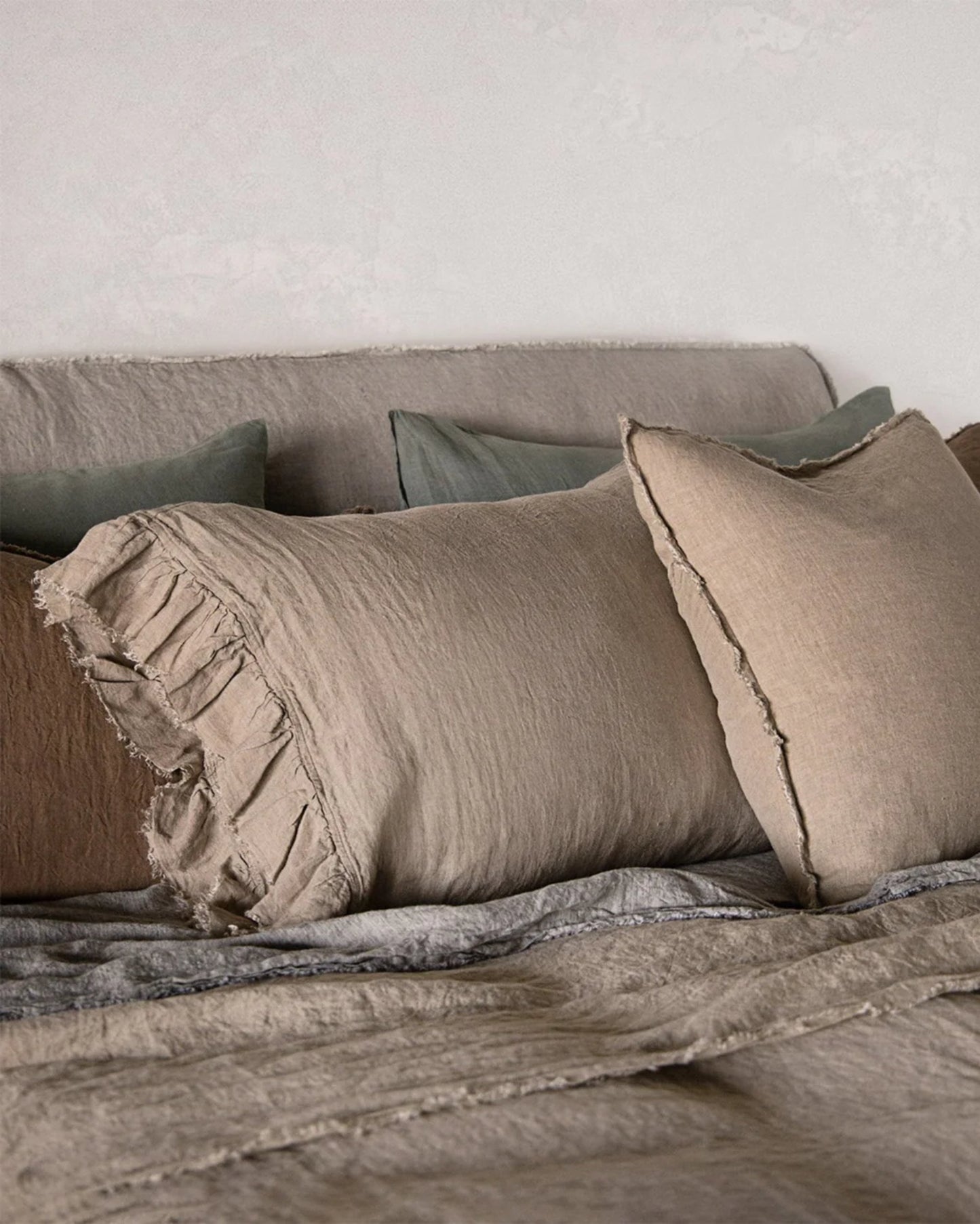 A cozy bed setup exudes rustic romance, featuring rumpled *Hale Mercantile Co HALE KRISTINE* standard pillowcases in beige and gray linen. Two beige pillows with fringed edges are prominently displayed, offering vintage appeal alongside darker cushions in the background, all set against a neutral wall.