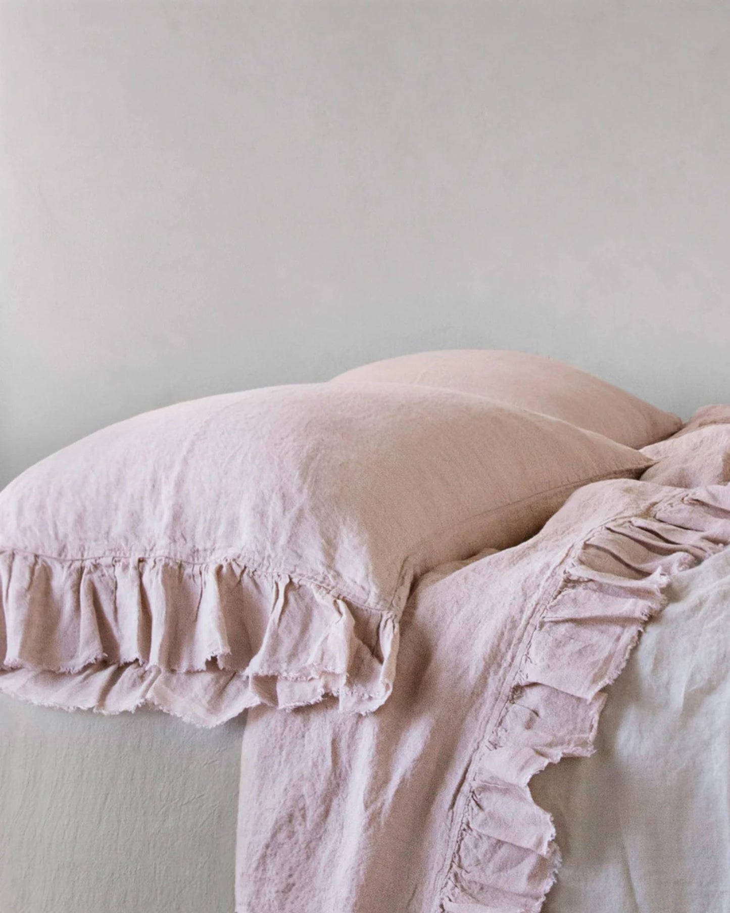 A neatly stacked set of HALE KRISTINE STANDARD PILLOWCASE in pale pink linen by Hale Mercantile Co against a soft, light-colored wall evokes rustic romance. The textured fabric enhances this cozy, inviting atmosphere with a touch of vintage appeal.