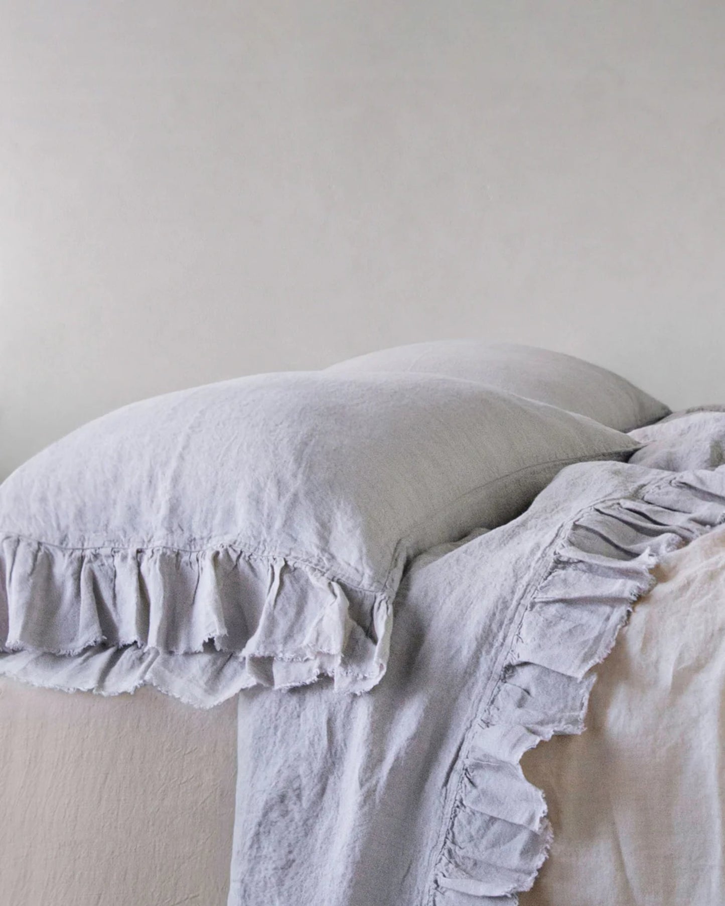 Against a plain light gray wall, a neatly made bed with two soft, pale gray HALE KRISTINE STANDARD PILLOWCASES from Hale Mercantile Co and a matching comforter, both featuring ruffled edges, exudes a vintage appeal.