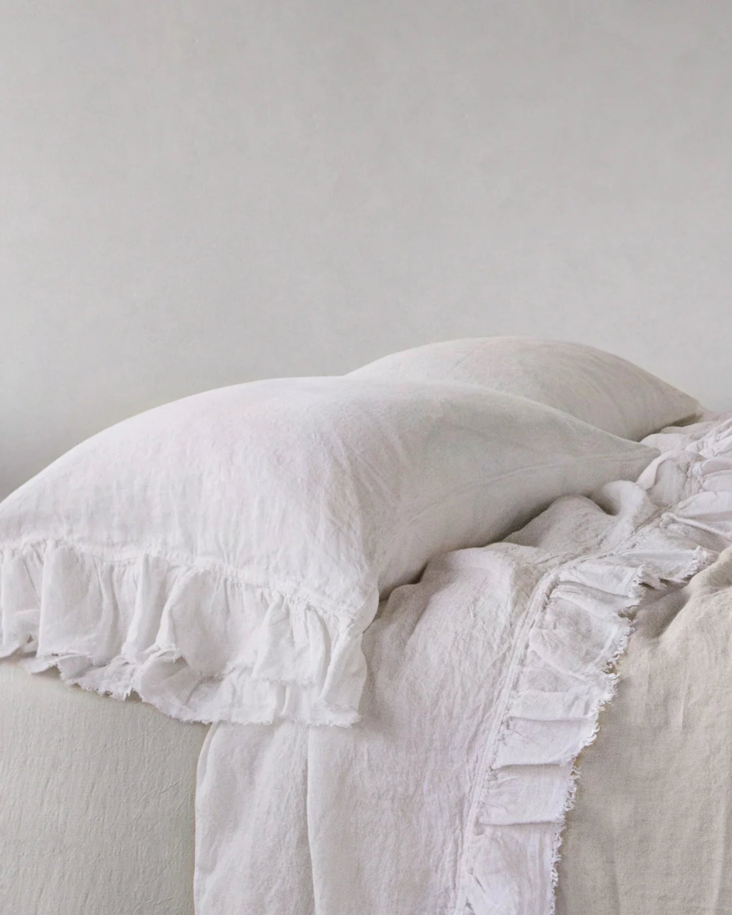 A neatly made bed showcases the HALE KRISTINE STANDARD PILLOWCASE by Hale Mercantile Co, featuring white linen pillowcases set against a light gray background. The bedding's vintage design, complete with soft textures and inviting appeal, creates a clean and cozy atmosphere.