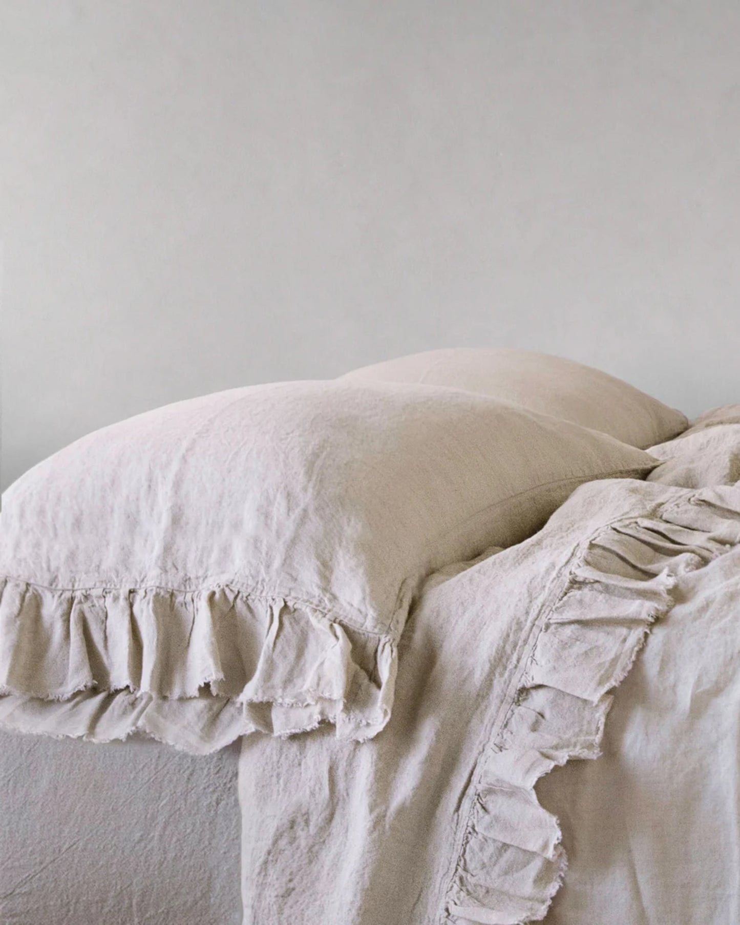 The cream-colored HALE KRISTINE STANDARD PILLOWCASES by Hale Mercantile Co, with ruffled detailing, and a matching blanket on a bed against a plain, neutral wall create a cozy and elegant atmosphere with vintage appeal.