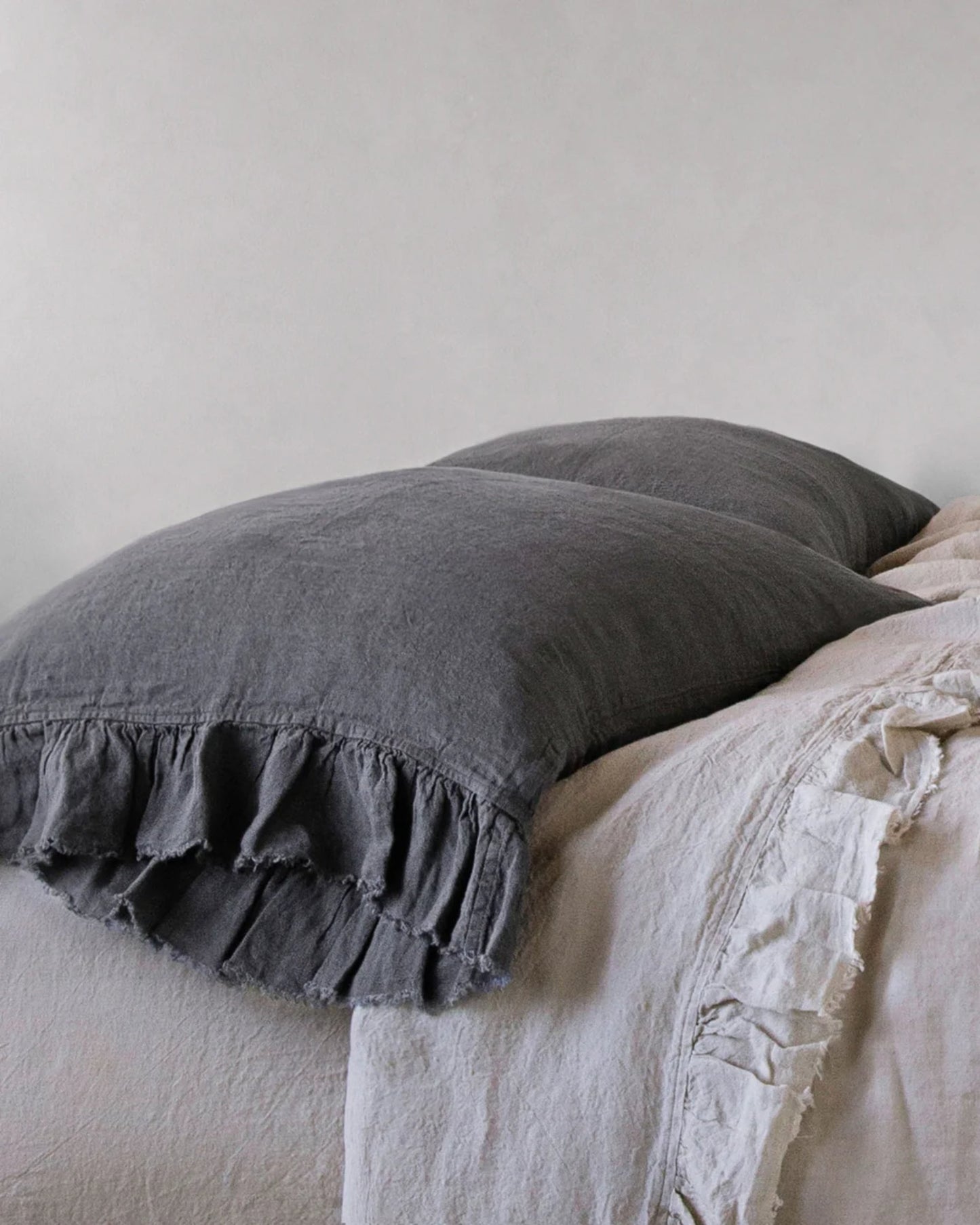 A neatly made bed showcases a dark gray pillow and a light gray ruffled blanket, exuding rustic romance. The HALE KRISTINE STANDARD PILLOWCASE by Hale Mercantile Co adds a touch of vintage appeal, enhancing the calm, neutral palette.