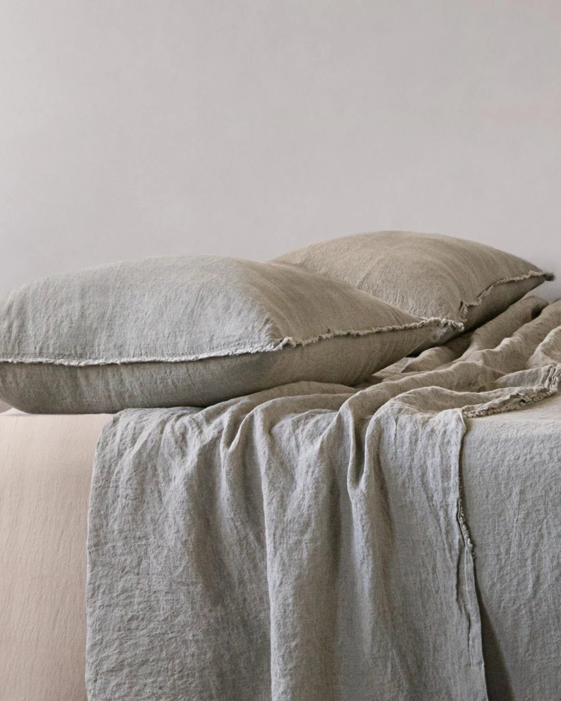 Close-up of a neatly made bed featuring soft, pure linen sheets and a plump, handcrafted pillow atop Hale Mercantile Co's HALE STANDARD FLOCCA PILLOWCASE. The sheets' gentle, wrinkled texture enhances the cozy and inviting atmosphere against the plain background.