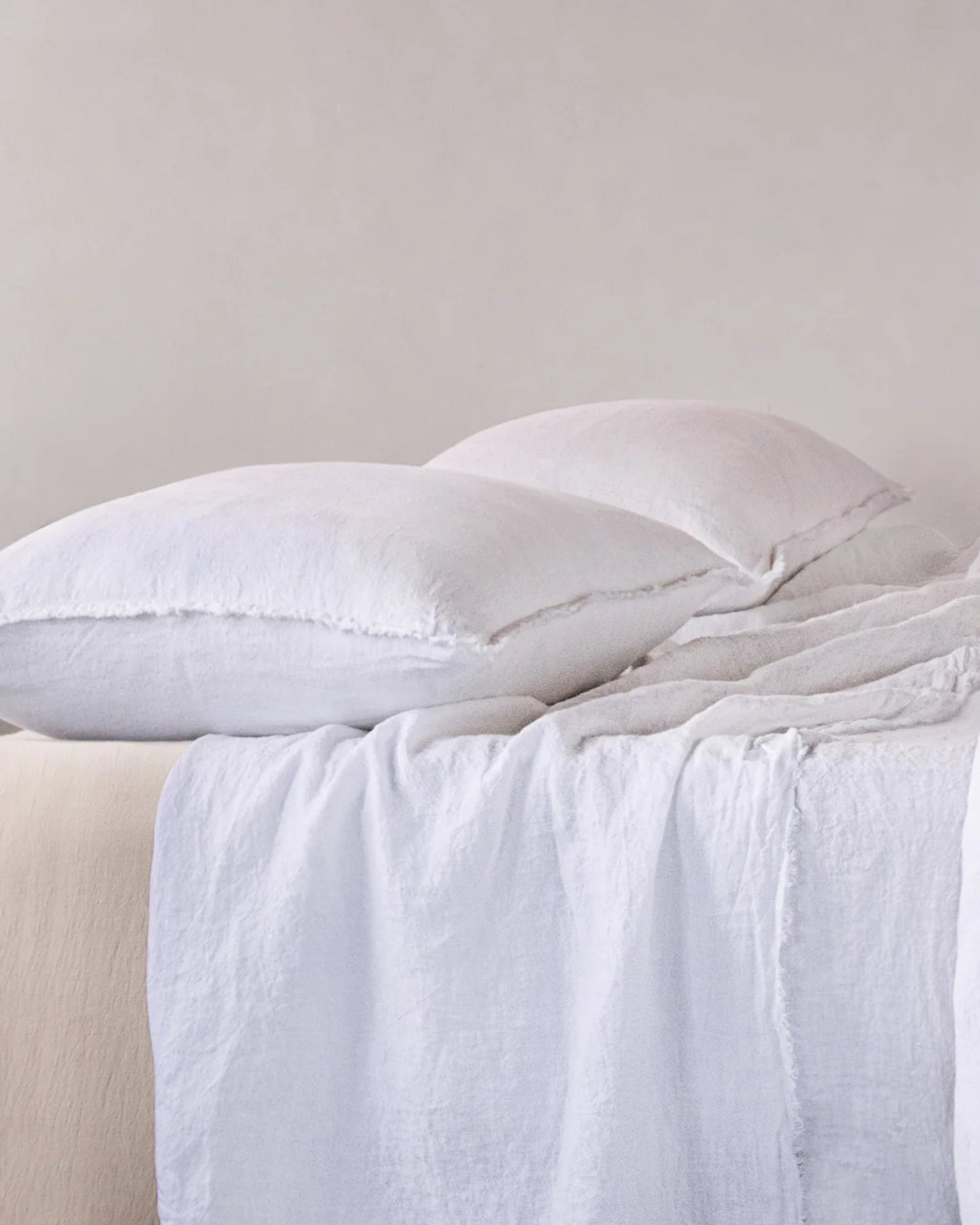 A neatly made bed showcasing two fluffy white pillows encased in HALE STANDARD FLOCCA PILLOWCASE by Hale Mercantile Co, complemented by a textured white blanket draped over the edge against a neutral background. The handcrafted setting conveys a sense of comfort and simplicity.