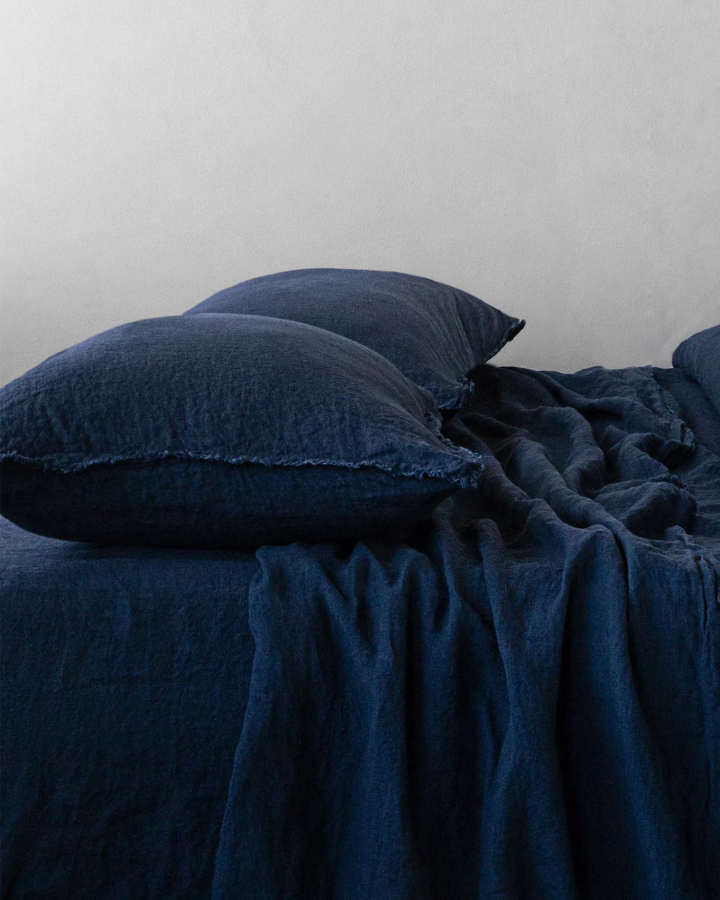 A neatly made bed features dark blue pure linen sheets and Hale Mercantile Co's HALE STANDARD Flocca Pillowcases against a plain white wall. The soft, textured appearance of the fabric creates a cozy and inviting atmosphere.