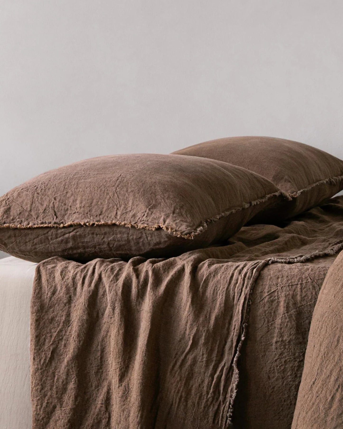 A neatly arranged bed showcases two large, handcrafted HALE STANDARD FLOCCA PILLOWCASES from Hale Mercantile Co, made of pure linen, accompanied by a matching crumpled brown blanket that creates a cozy and inviting atmosphere. The background is a neutral off-white wall.