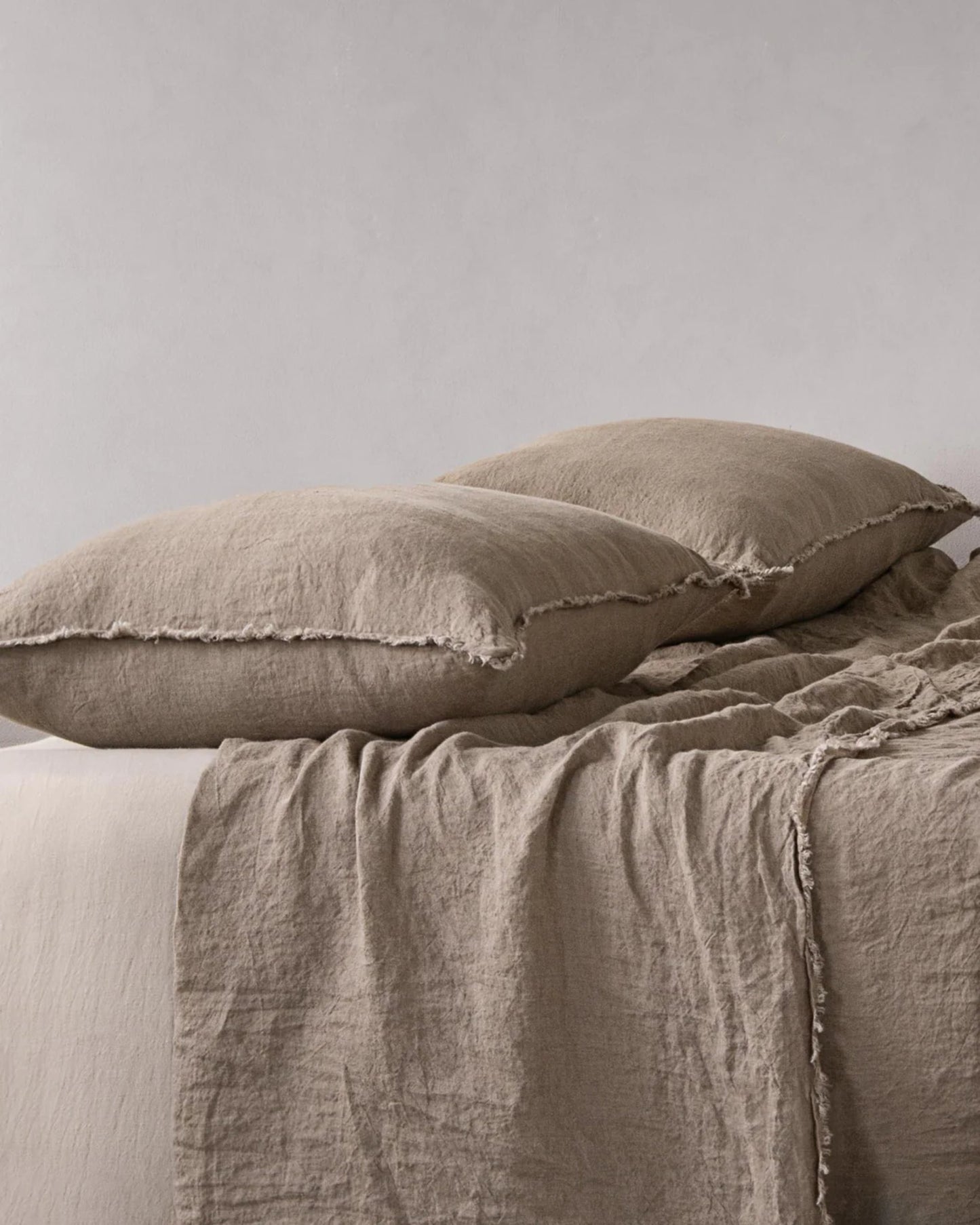 A neatly made bed showcases two large, soft beige Hale Standard Flocca Pillowcases by Hale Mercantile Co, paired with a matching pure linen sheet, establishing a cozy and minimalist aesthetic against a plain light gray background.