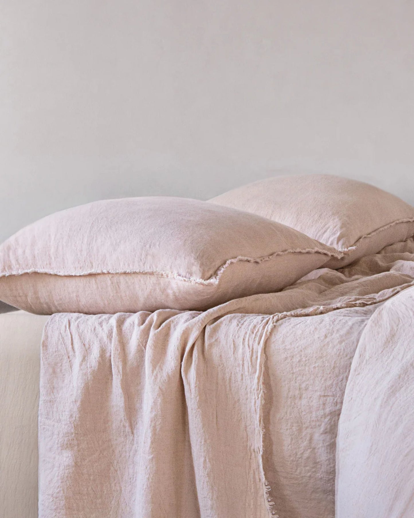 A neatly arranged bed showcases soft, light pink Hale Mercantile Co HALE STANDARD FLOCCA PILLOWCASES alongside a matching linen duvet. The bedding's slightly crumpled texture contributes to a cozy, inviting ambiance in the light-colored room, further enhanced by the allure of handcrafted pillowcases.