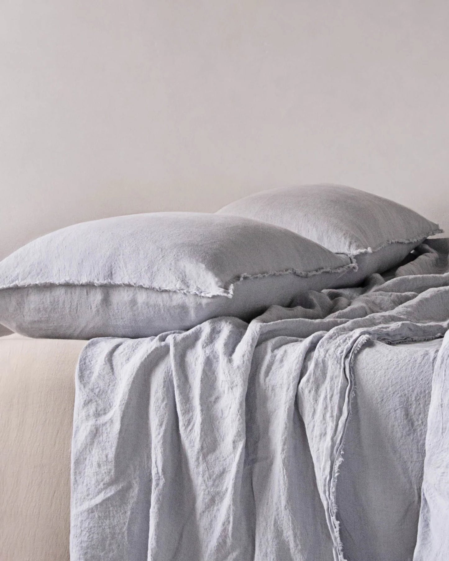 A neatly made bed showcases the HALE STANDARD FLOCCA PILLOWCASE by Hale Mercantile Co, paired with a matching draped bedding set in light gray. The overall aesthetic is soft and minimalistic, creating a serene and comfortable ambiance in this neutral-toned bedroom featuring pure linen.