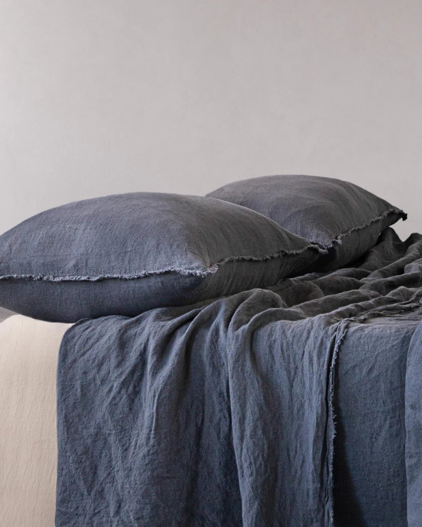 A bed featuring a dark gray pure linen duvet cover and HALE STANDARD FLOCCA PILLOWCASE by Hale Mercantile Co, set against a plain light gray wall. The fabric's soft, slightly wrinkled texture imparts a casual and cozy appearance.