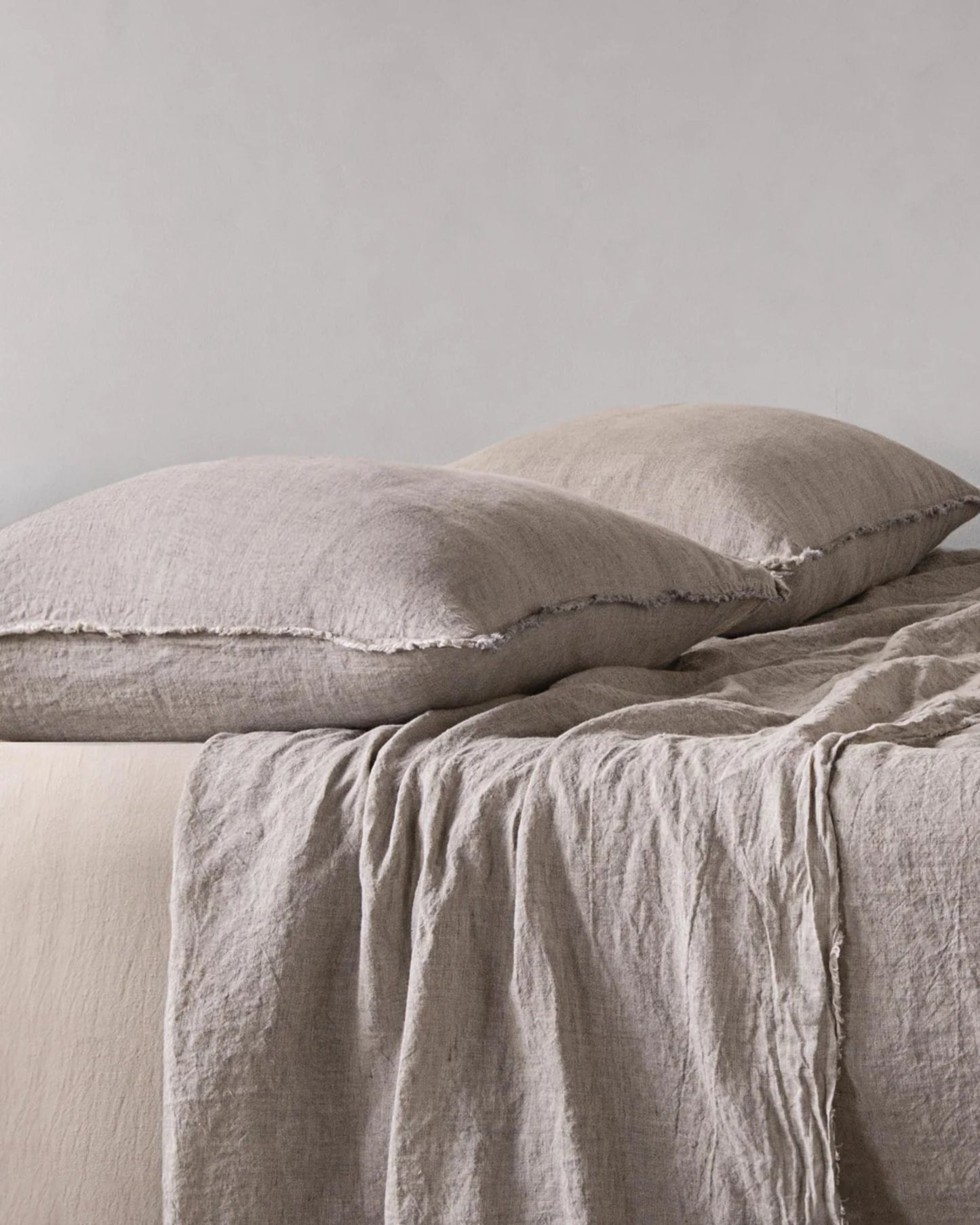 A neatly made bed features two large HALE STANDARD FLOCCA PILLOWCASES by Hale Mercantile Co and a matching beige duvet cover draped over the mattress. The background is a plain, soft gray wall, cultivating a calm and minimalist bedroom setting.