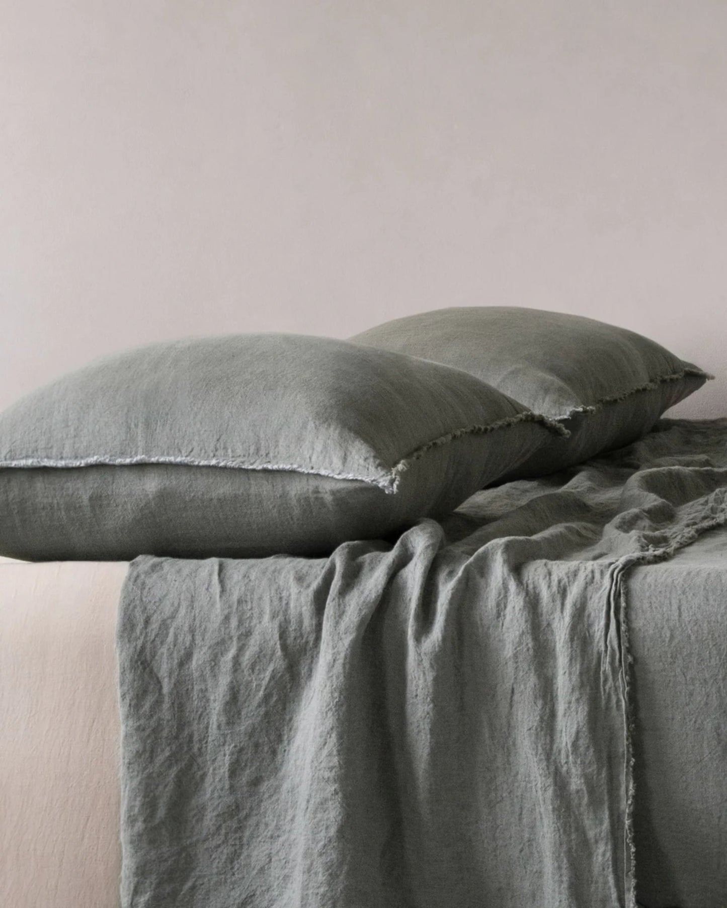 Two soft HALE STANDARD FLOCCA PILLOWCASES in light gray are placed on a bed adorned with a complementary textured gray blanket. This bedding, crafted from pure linen by Hale Mercantile Co, boasts a casual and slightly wrinkled look that enhances the cozy and relaxed ambiance of the room.