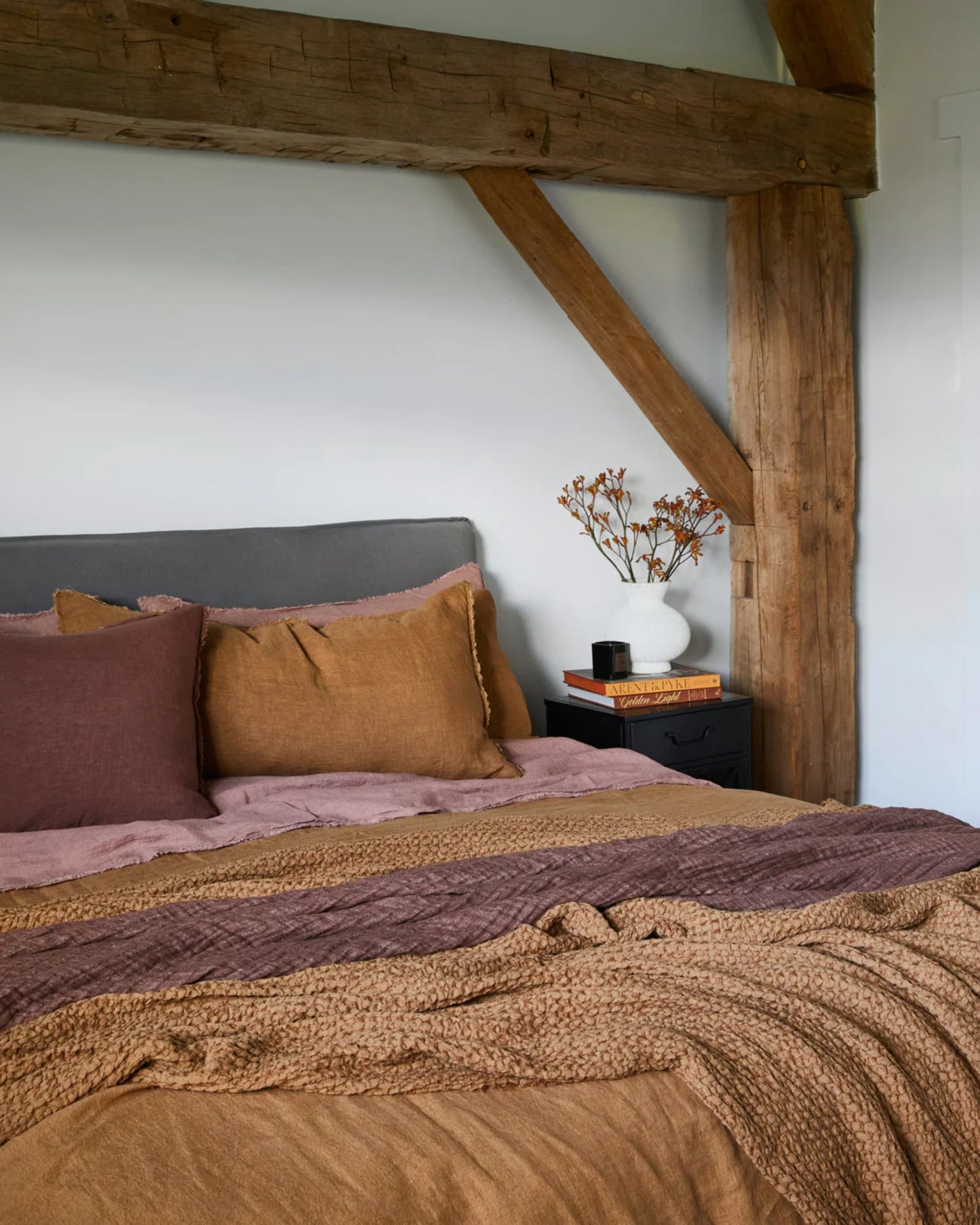 A cozy bedroom showcases a large wooden beam and a neatly made bed adorned with earth-toned linens and a gray headboard. On the nightstand, you'll find a stack of books, a small potted plant, and Hale Mercantile Co's HALE STANDARD FLOCCA PILLOWCASE, adding to the inviting atmosphere with their comforting touch.