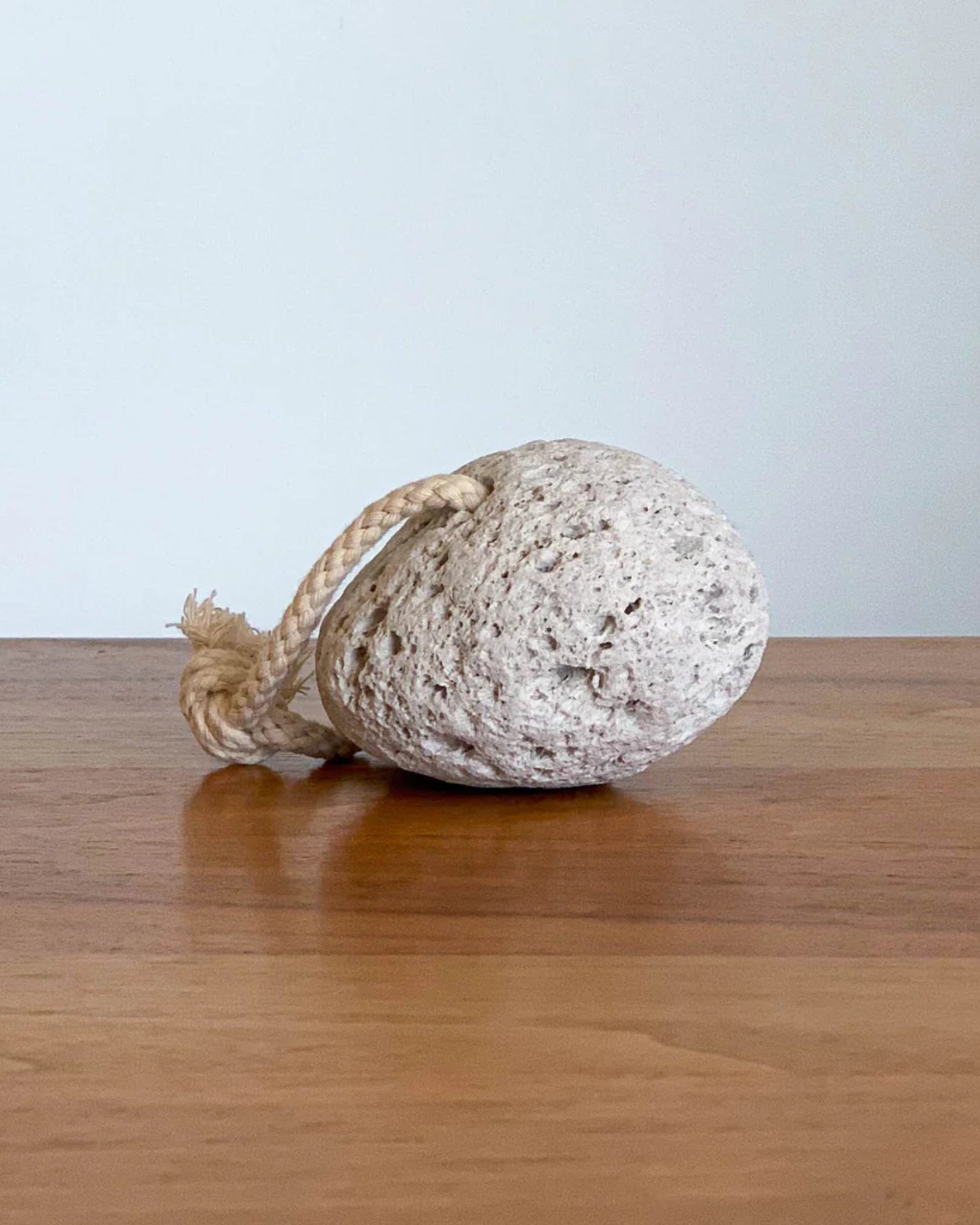 The HAMMAN PUMICE STONE by Redecker, featuring a rough, spherical design with a short rope loop attached, rests on a wooden surface. Against a plain, light-colored wall backdrop, its texture is highlighted, promising to leave your skin soft and smooth by effectively removing dry skin.