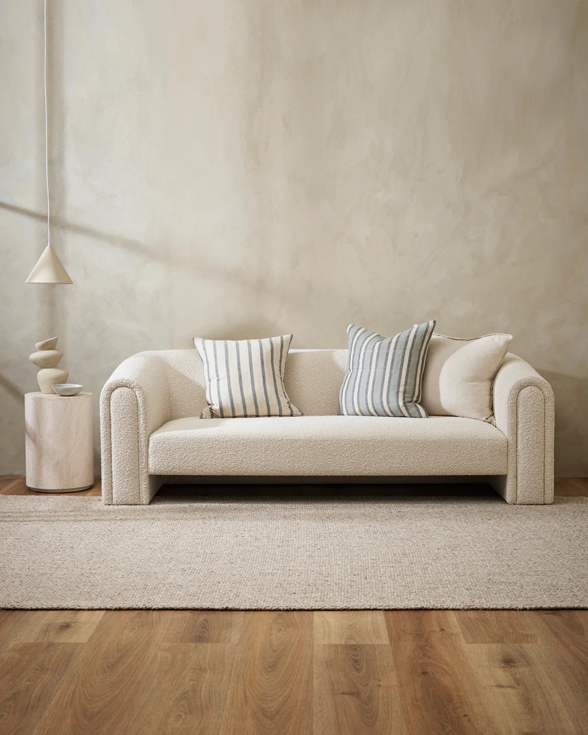 A minimalist living room is furnished with a cream-colored sofa accentuated by Baya's HAMPTON CUSHION, featuring a casual stripe design. It includes a textured area rug made of natural fibers and a small side table adorned with abstract decor. Soft lighting and neutral tones contribute to a serene and cozy ambiance.