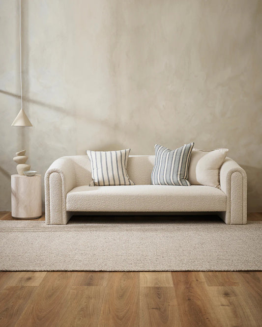 A minimalist living room is furnished with a cream-colored sofa accentuated by Baya's HAMPTON CUSHION, featuring a casual stripe design. It includes a textured area rug made of natural fibers and a small side table adorned with abstract decor. Soft lighting and neutral tones contribute to a serene and cozy ambiance.