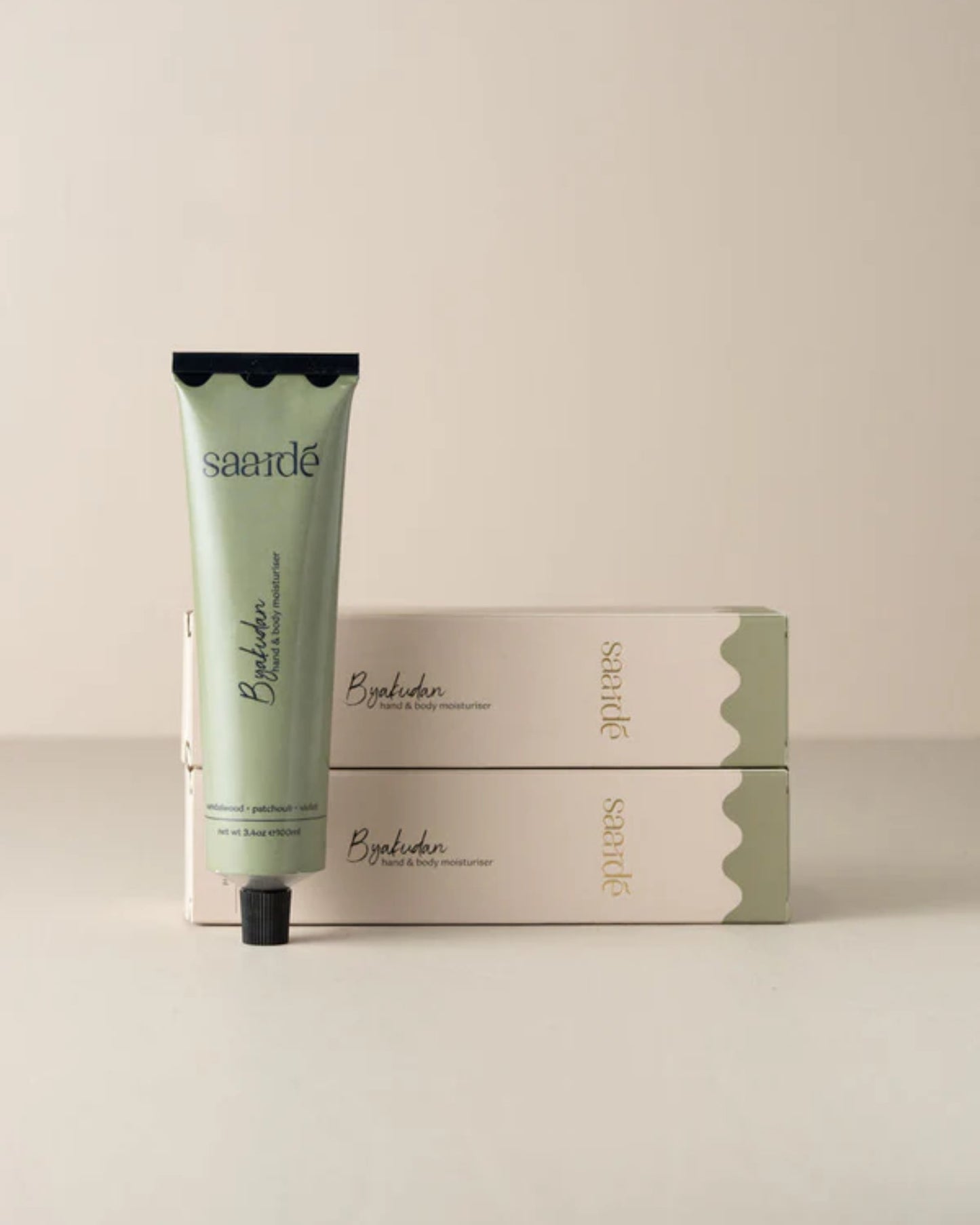 A tube of Saarde's HAND AND BODY MOISTURISER in green, enriched with extra virgin olive oil, stands upright before two stacked boxes against a neutral background, featuring simple and elegant packaging.