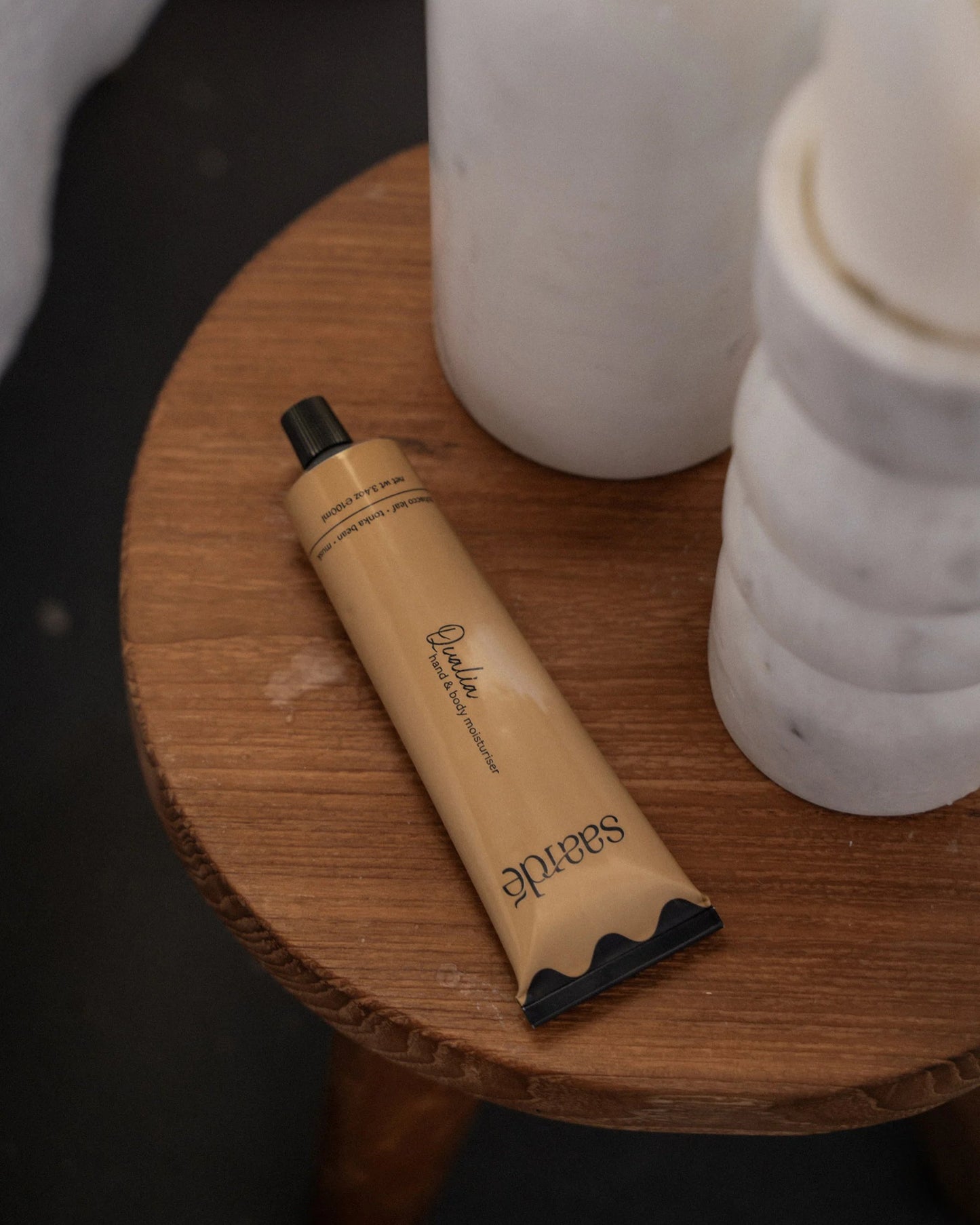 A tube of Saarde HAND AND BODY MOISTURISER featuring black detailing rests on a wooden surface beside three white rolled-up towels, enhanced with the calming essence of sandalwood. The scene exudes a minimalist and natural aesthetic.