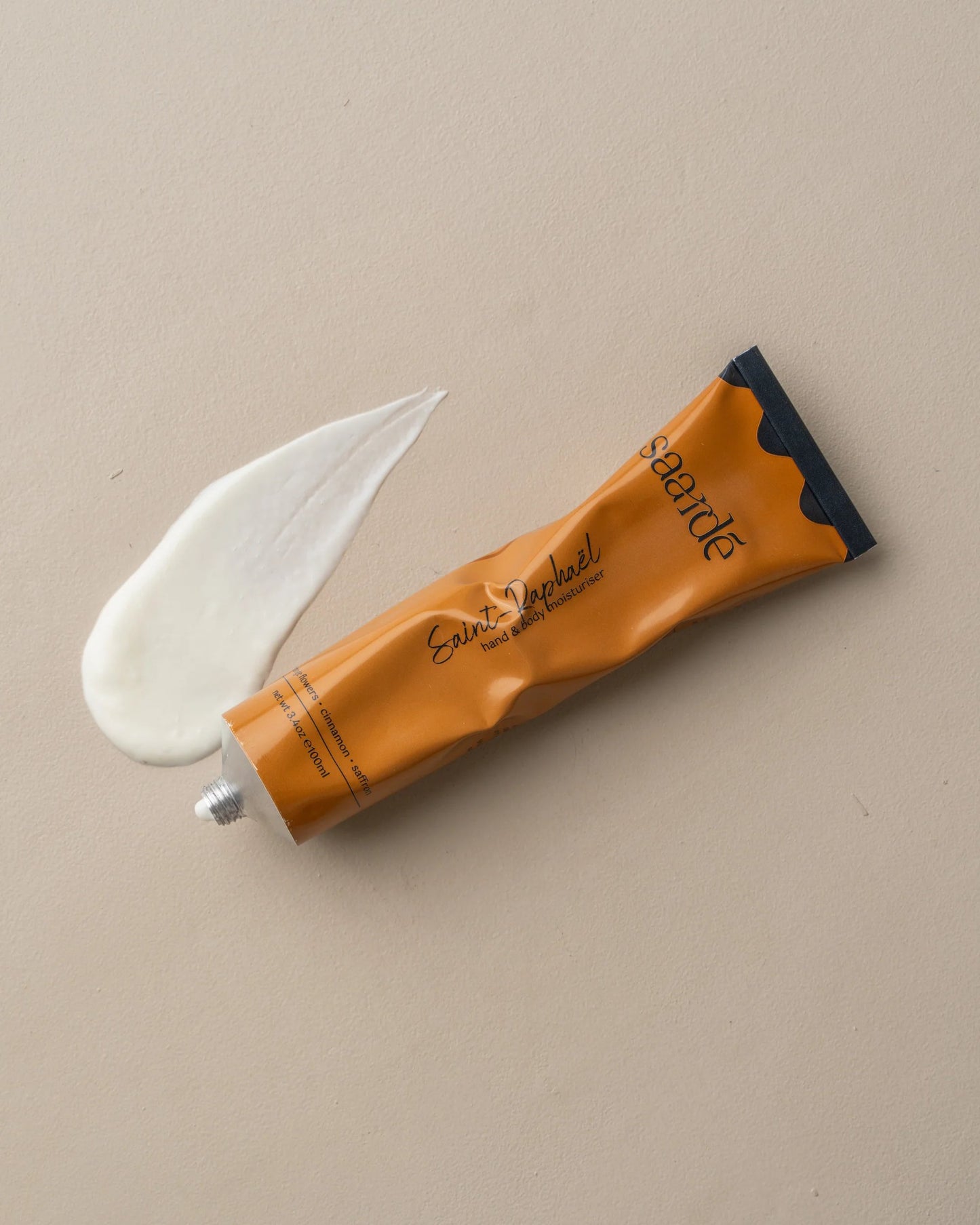 A tube of Saarde's HAND AND BODY MOISTURISER in orange and black rests on a beige surface. The open tube reveals a small amount of creamy white lotion, enriched with sandalwood, spread next to it—ideal for achieving silky smooth skin.