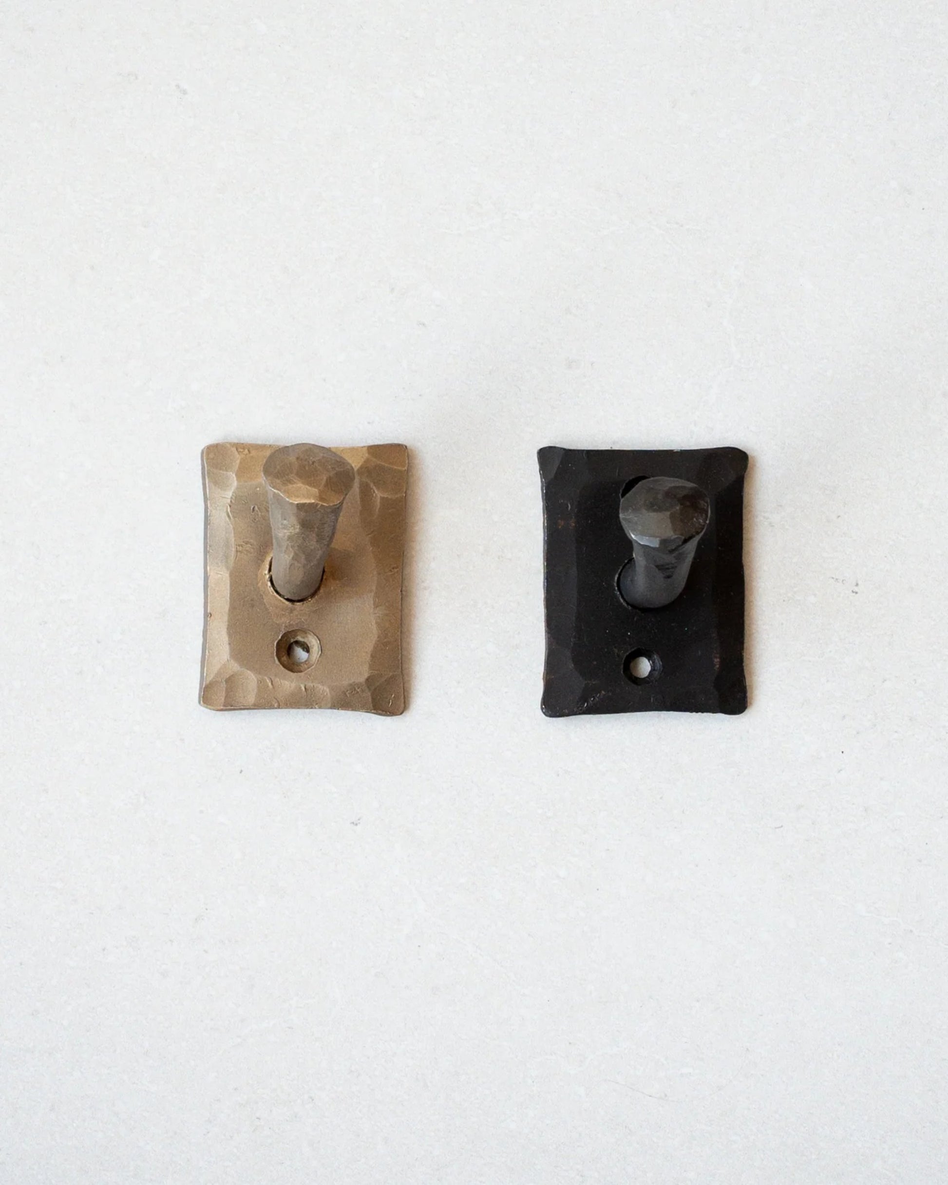 On a light-colored wall, two rectangular hooks from New Direction are displayed. The left hook features a hand-forged finish in brass with a textured touch. The right hook, in dark gray, reflects a rustic industrial style. Both HANDMADE NAIL HOOKs exhibit a simple and functional design that embodies the charm of handmade iron nails.
