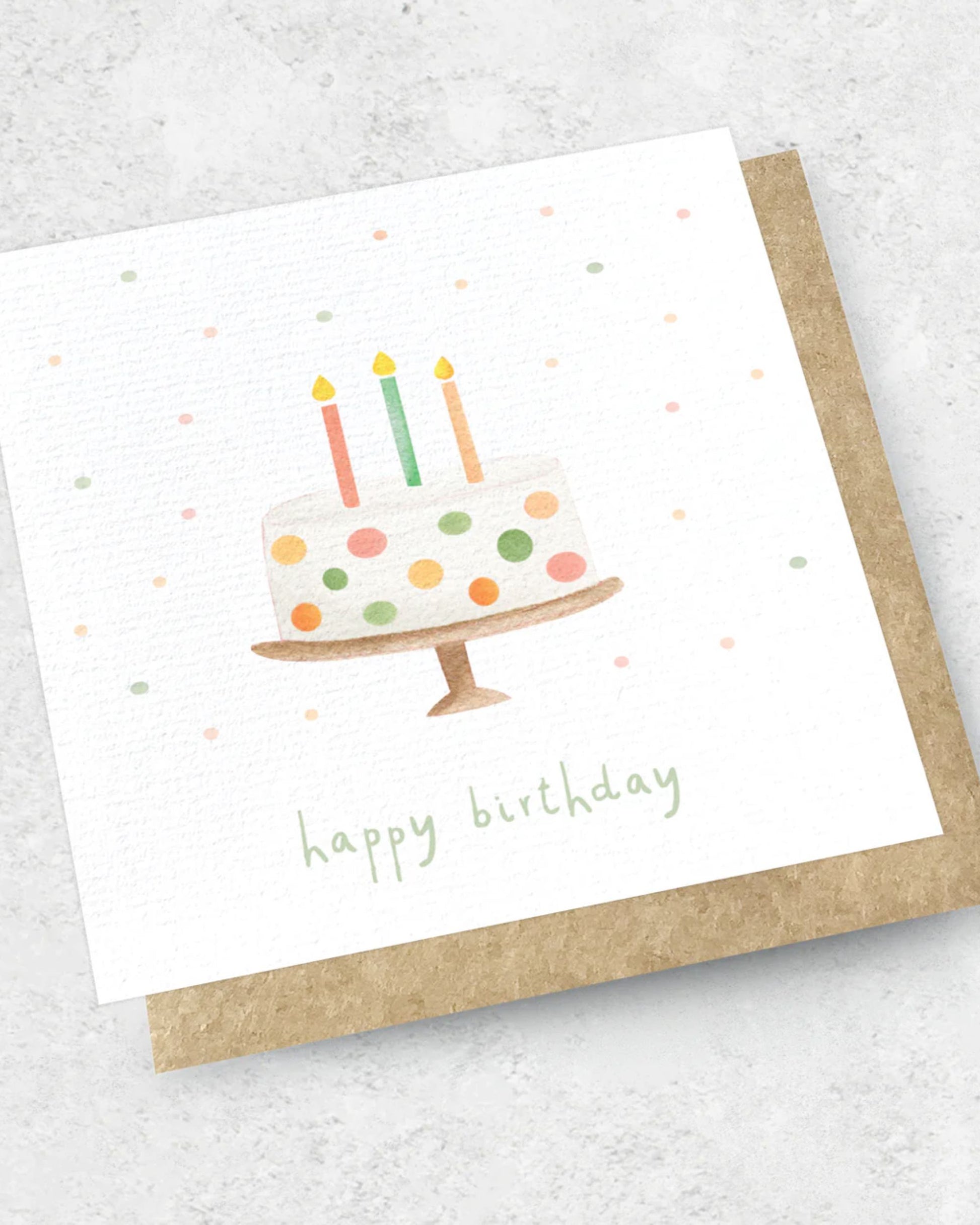 Introducing the HAPPY BIRTHDAY CAKE - GREETING CARD by Ink Bomb: This exquisite card showcases a watercolor illustration of a birthday cake on a stand, complete with three colorful candles and delightful dots. The words "Happy Birthday" are gracefully inscribed in green text beneath the illustration. Paired with a kraft envelope, this premium textured card adds an extra touch of charm to any celebration.