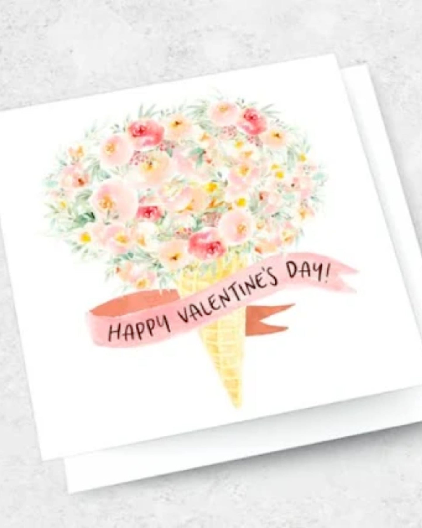 The HAPPY VALENTINES DAY - FLORAL ICE CREAM GREETING CARD showcases a bouquet of pink and peach flowers creatively arranged in the shape of an ice cream cone, complete with a "Happy Valentines Day!" banner. This premium textured card comes with a matching kraft envelope, all set against a light, textured background.