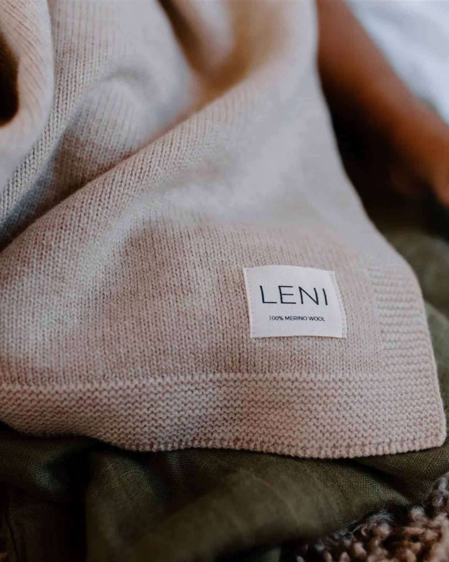 A close-up of a beige HEIRLOOM MERINO BLANKET showcases a label with "Leni 100% Merino Wool" sewn onto it. Known for its breathability and ability to regulate temperature, the fabric is elegantly draped over what seems to be green material.