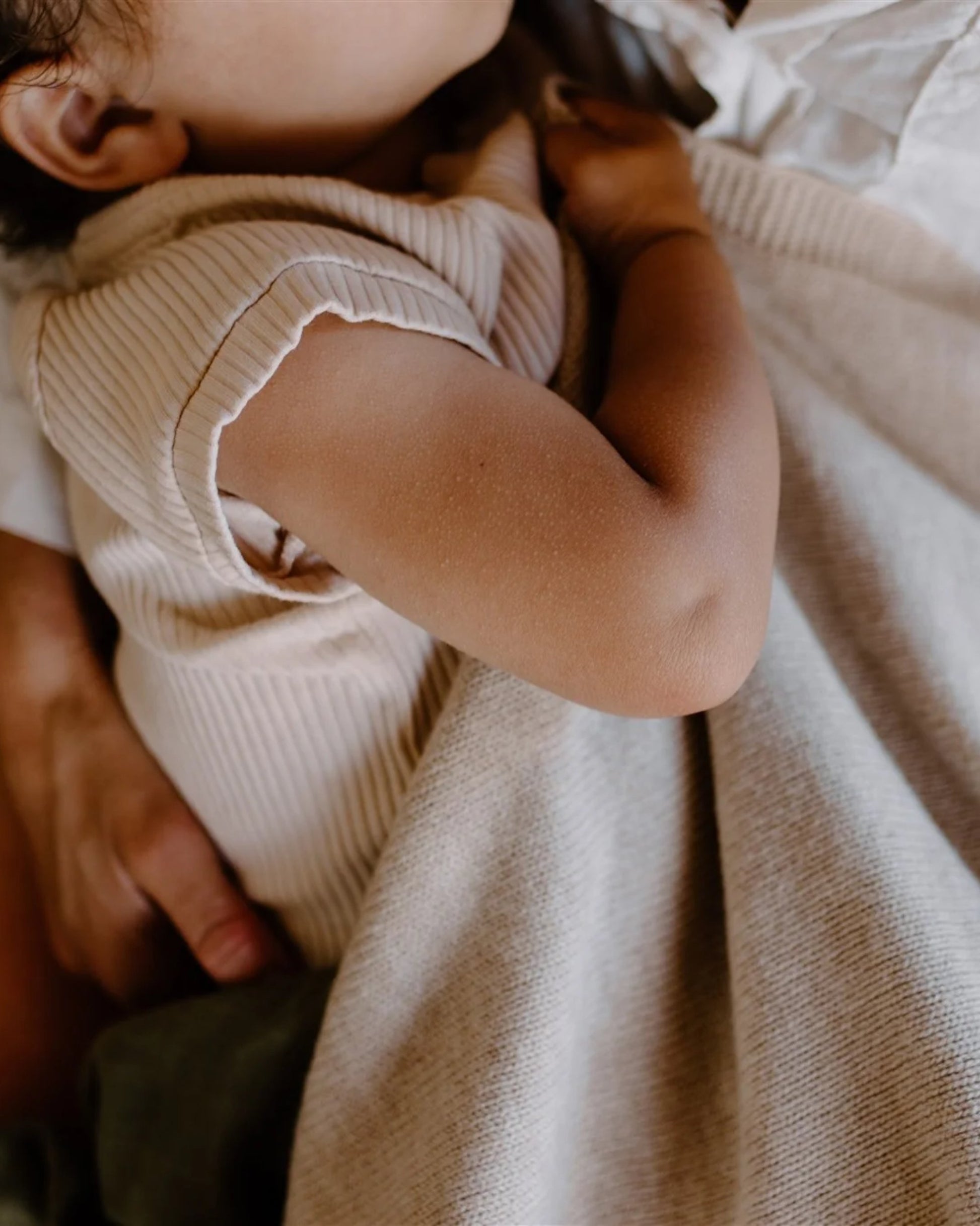 A close-up shows a baby resting peacefully, dressed in a beige ribbed outfit. The baby is gently held in an adult's arms against Leni's HEIRLOOM MERINO BLANKET, renowned for its temperature regulation and breathability, creating a tender and serene atmosphere.