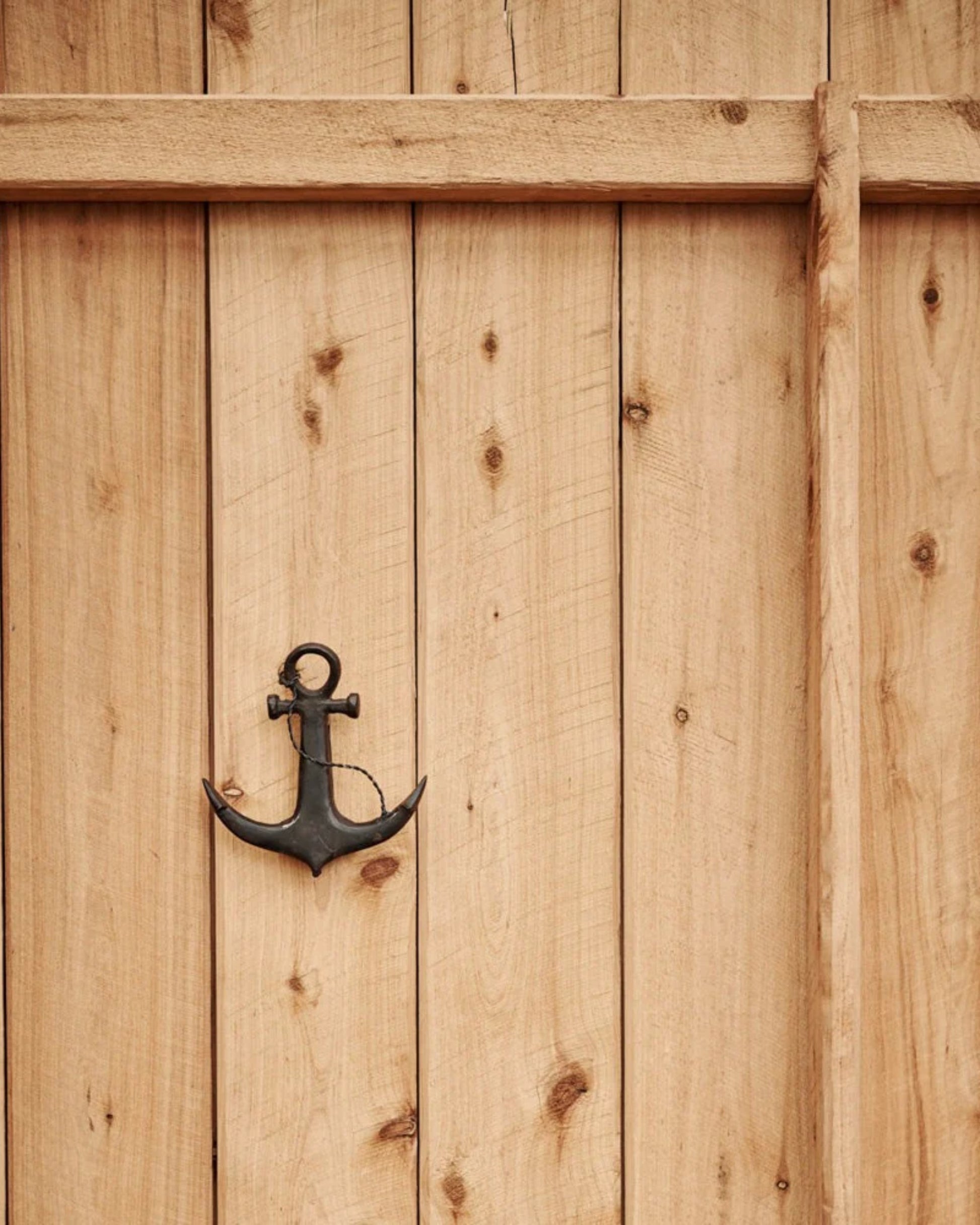 A wooden fence adorned with vertical planks is enhanced by a decorative metal anchor and the Society Ink HELLO SAILOR KNOCKER, bringing a nautical flair to your home decor.