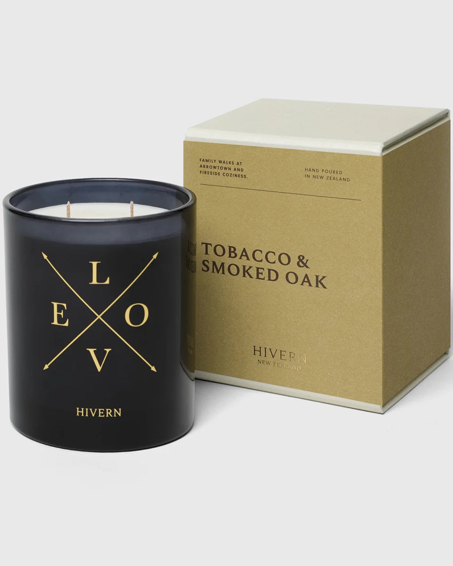 A black HIVERN CANDLE featuring the word LOVE in intersecting gold letters is placed beside a beige and black box labeled TOBACCO & SMOKED OAK, designed by the New Zealand home fragrance brand Hivern. With a burn time of 55 hours, it offers lasting warmth and sophistication.