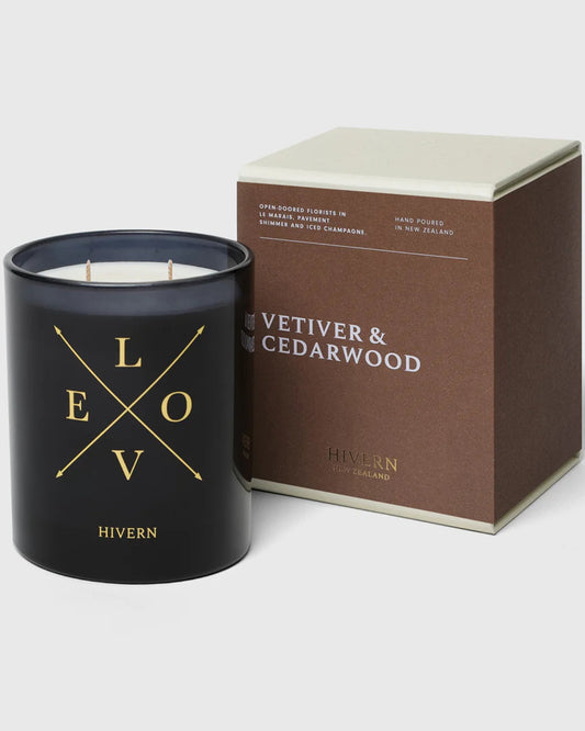 A black candle jar by Hivern, adorned with the word LOVE in gold lettering, is situated beside a brown and beige box labeled Vetiver & Cedarwood. This HIVERN CANDLE, a signature of New Zealand home fragrance, offers an impressive burn-time of 55 hours.