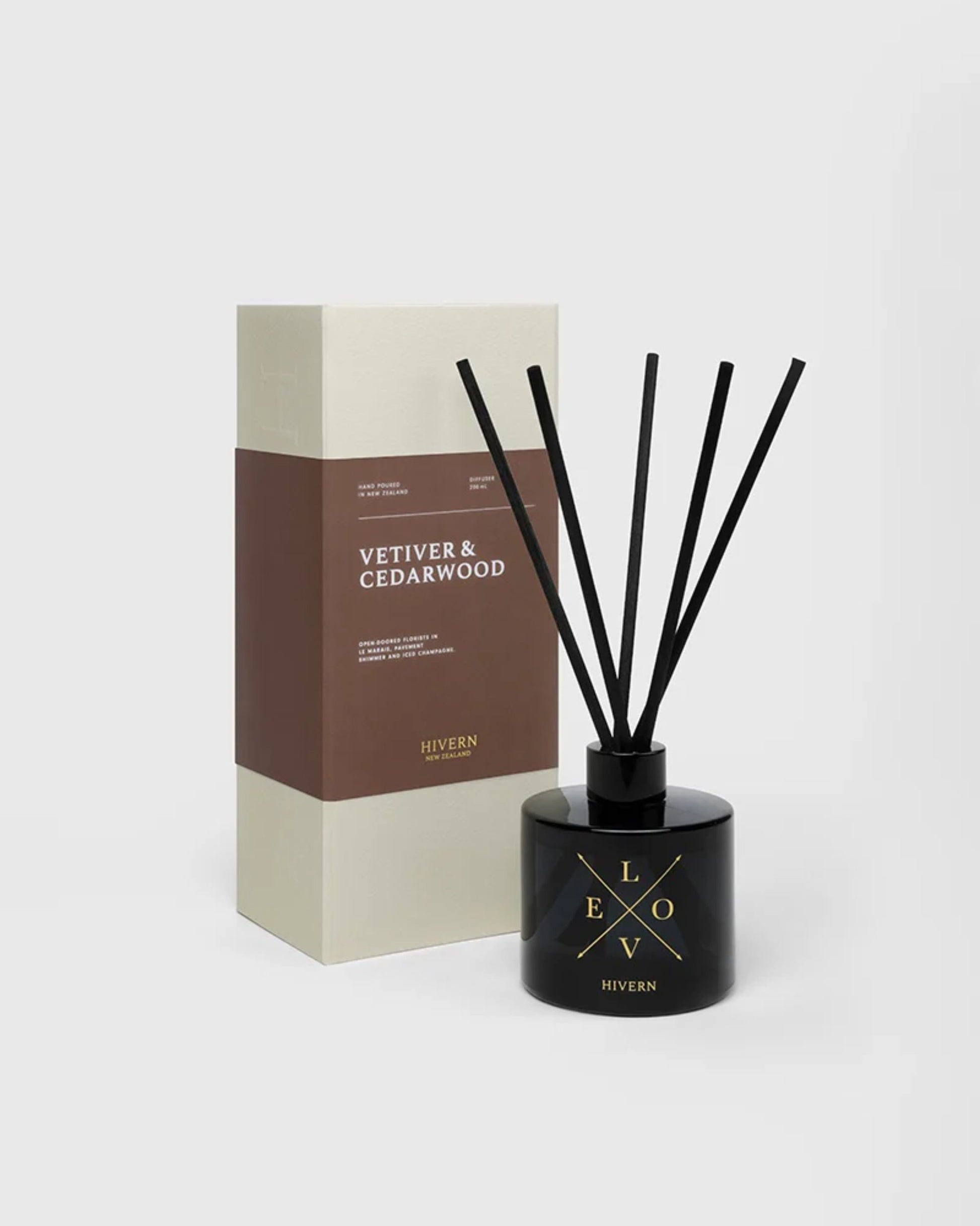 A black HIVERN DIFFUSER from Hivern, complete with five black silk fiber reeds, is placed next to a minimalist brown and cream box labeled Vetiver & Cedarwood. Both pieces are tastefully arranged against a plain gray background, exuding an air of understated luxury and optimal scent distribution.