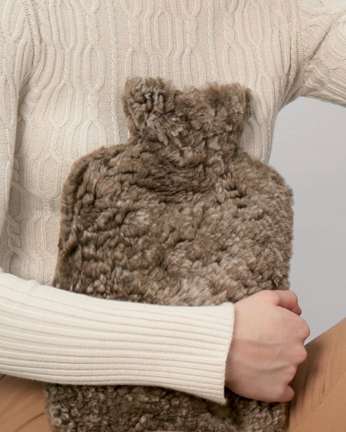 A person in a cream-colored cable knit sweater embraces the Bianca Lorenne NEW ZEALAND SHORT WOOL HOT WATER BOTTLE, featuring a luxurious sheepskin cover. The neutral background enhances the cozy, comforting feel of this indulgent scene.