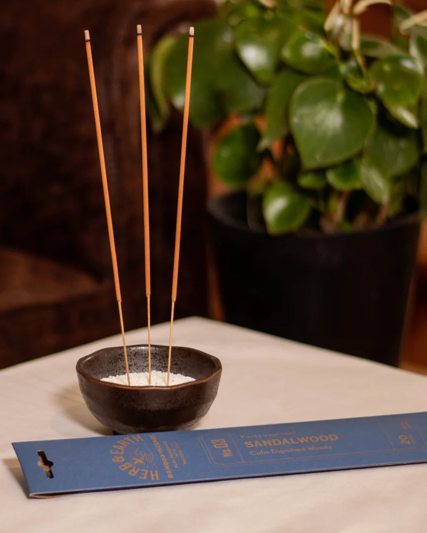 On a table, a small black bowl containing four bamboo incense sticks releases a pure fragrance with minimal smoke. Next to the bowl is blue packaging labeled "Herb and Earth - Sandalwood," while a lush green plant in a black pot provides the backdrop.