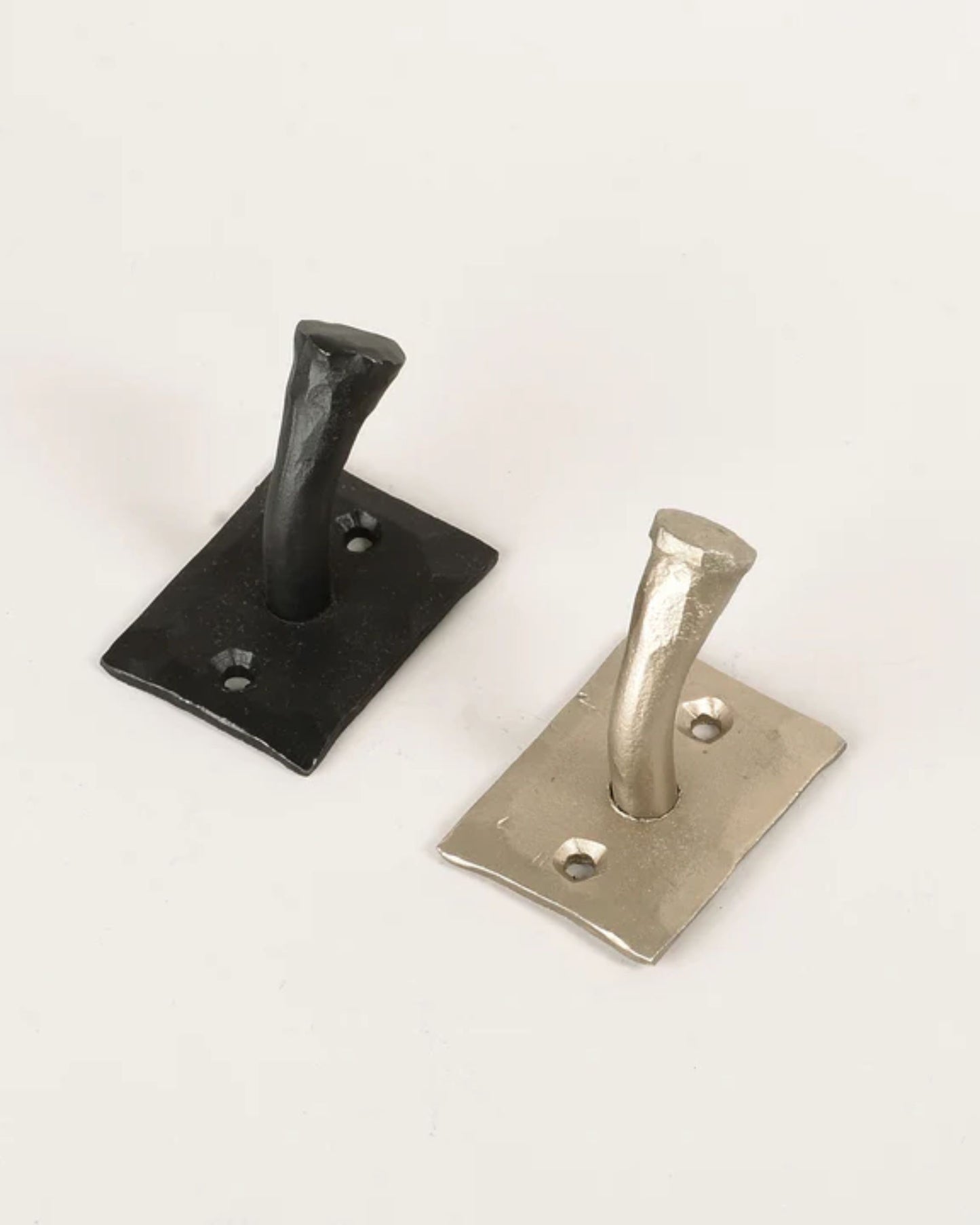 The HANDMADE NAIL HOOK by New Direction features a pair of hammered metal hooks designed with a rustic industrial style, mounted on rectangular backplates. Elegantly arranged on a light surface, one hook is in black while the other boasts a metallic silver finish. Each backplate includes two screw holes for straightforward installation, highlighting their hand-forged craftsmanship.