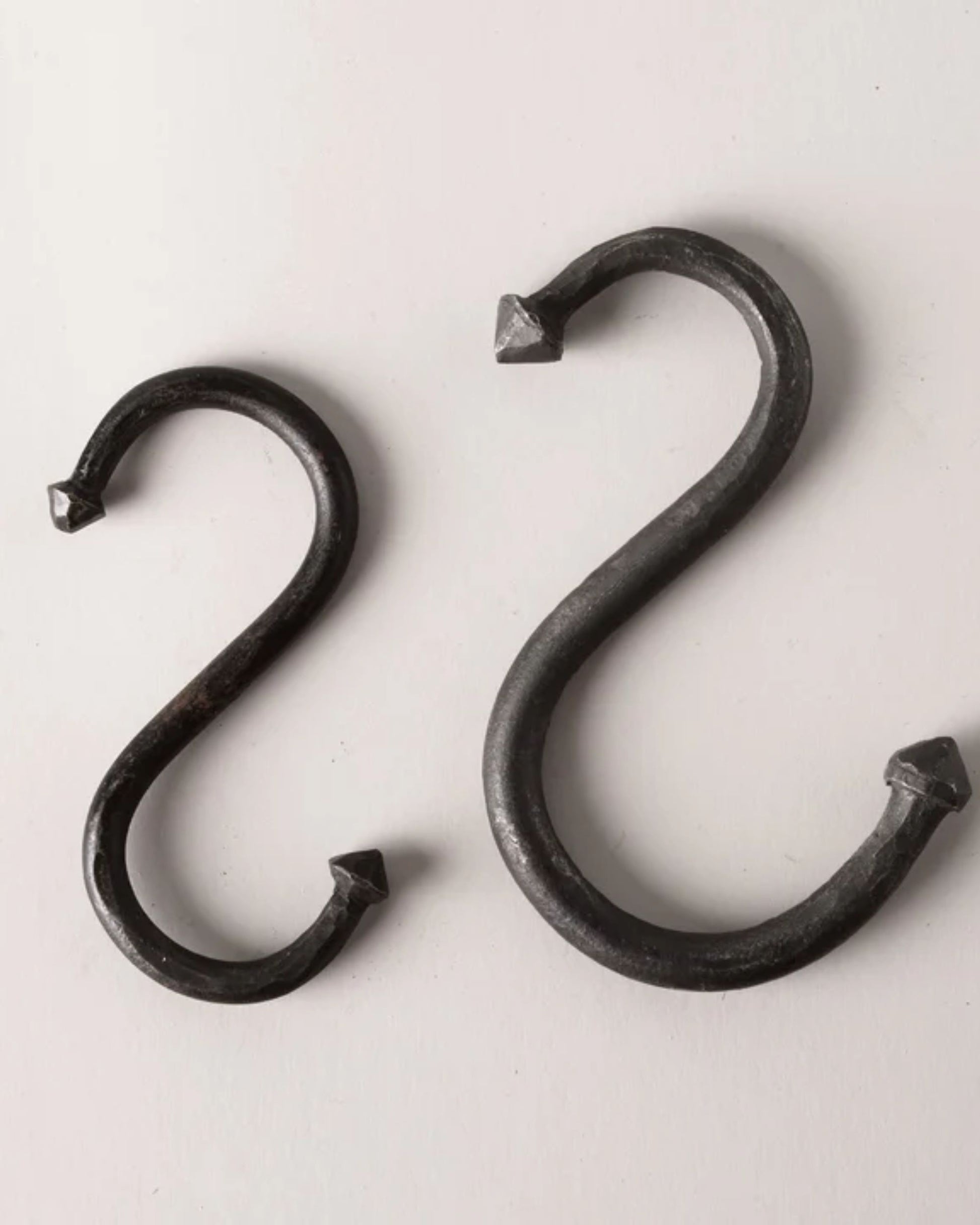 The IRON S HOOKS by New Direction, featuring two black metal hooks of varying sizes, are set against a light gray background. Designed from durable iron, they lend an industrial feel to any space. With pointed ends, these hooks provide a versatile storage solution for your home or workspace.