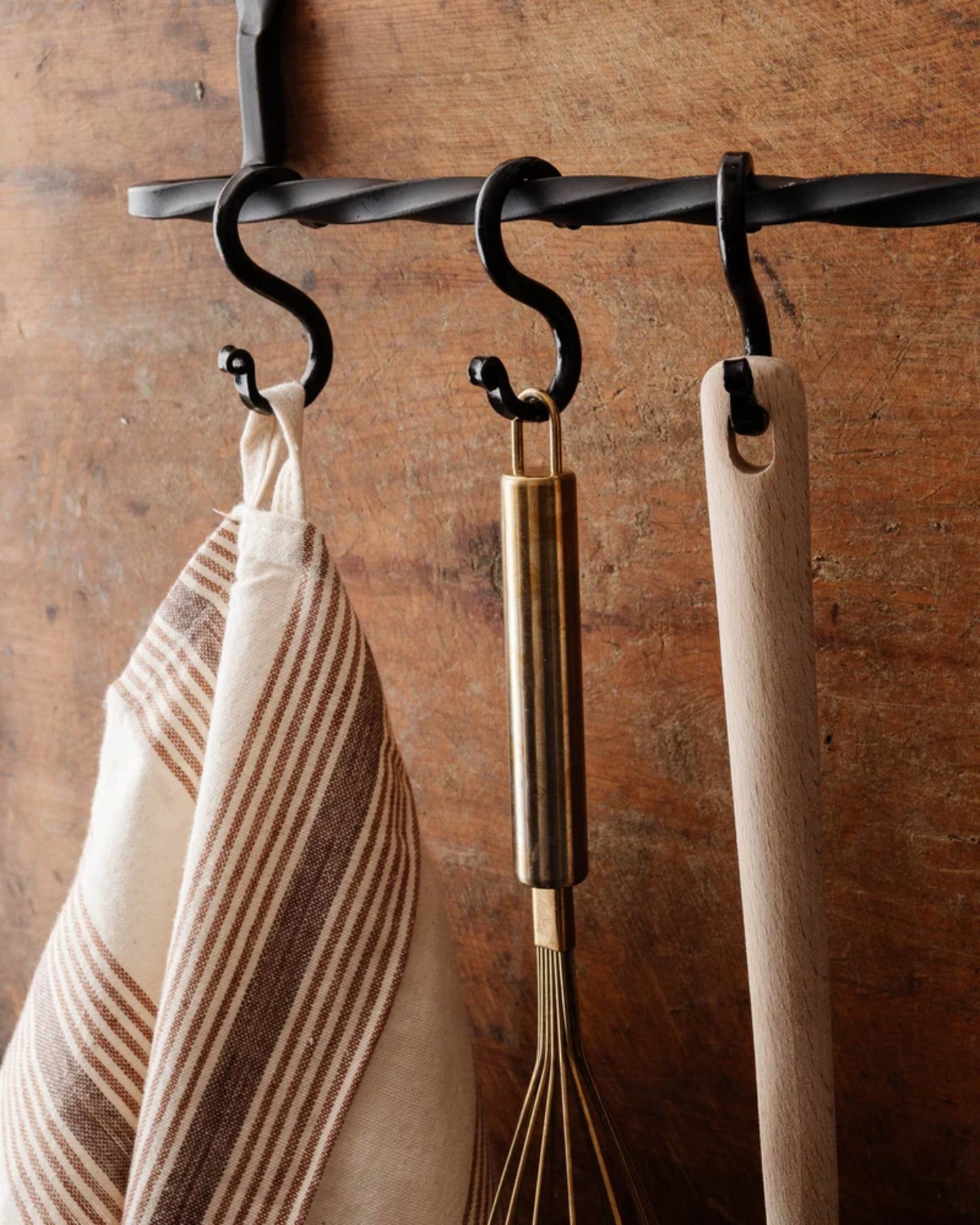 In a kitchen scene with an industrial flair, the New Direction IRON S HOOK offers a stylish storage solution. It elegantly showcases three items: a striped dish towel, a brass-colored whisk, and a wooden-handled utensil against the rustic charm of the wooden backdrop.