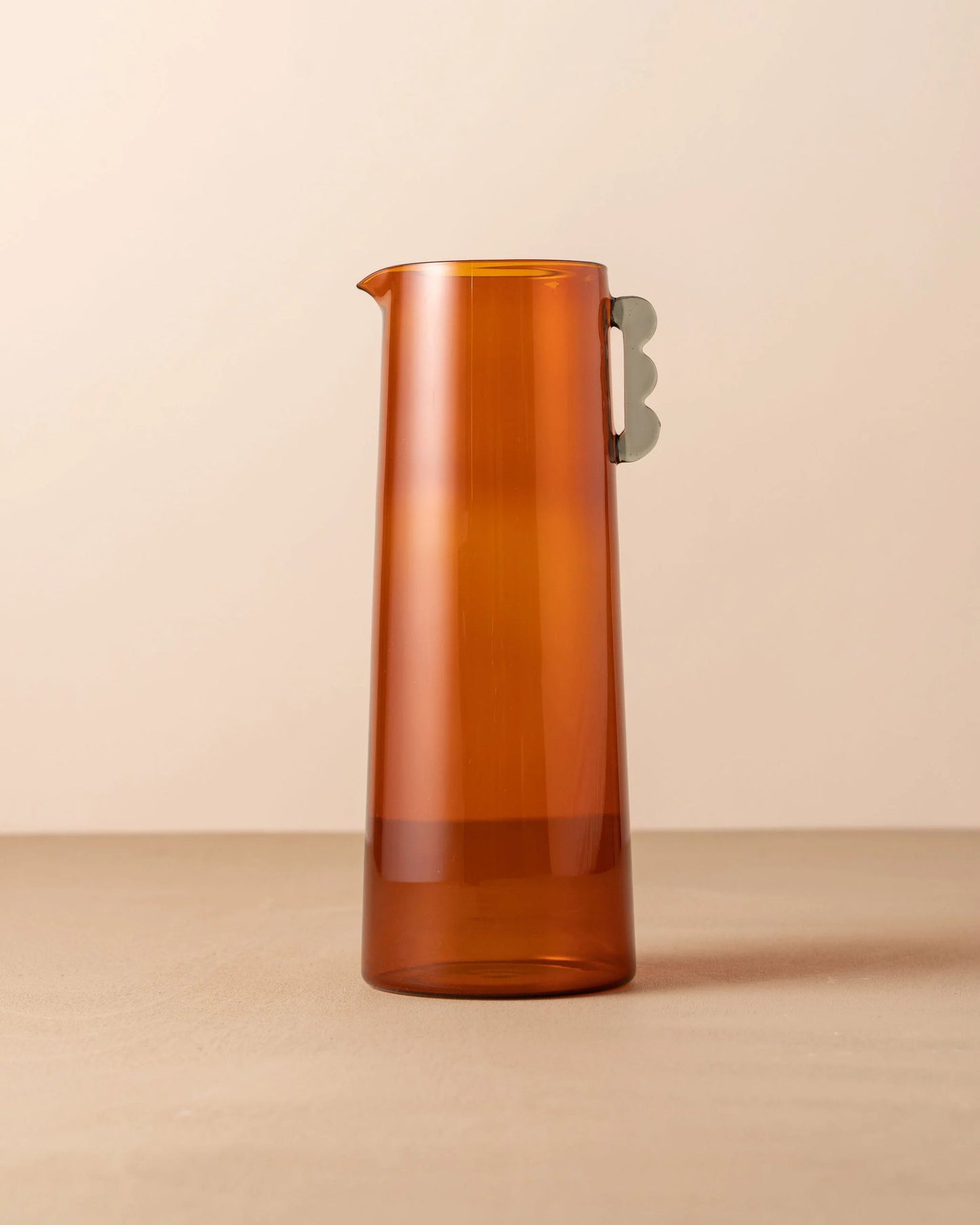 The KAIROS CARAFE by Saarde, a tall and sleek amber glass pitcher with a distinctive silver handle, reminiscent of Italian glassware, stands gracefully on a neutral surface against a beige background.