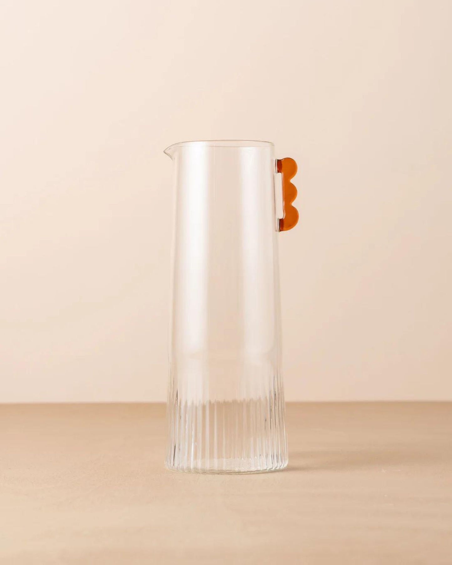 The KAIROS CARAFE by Saarde is a tall, clear glass pitcher made from borosilicate glass, showcasing a ribbed design on the lower half and highlighted with a scalloped orange handle. This elegant piece is set against a plain, soft beige background.