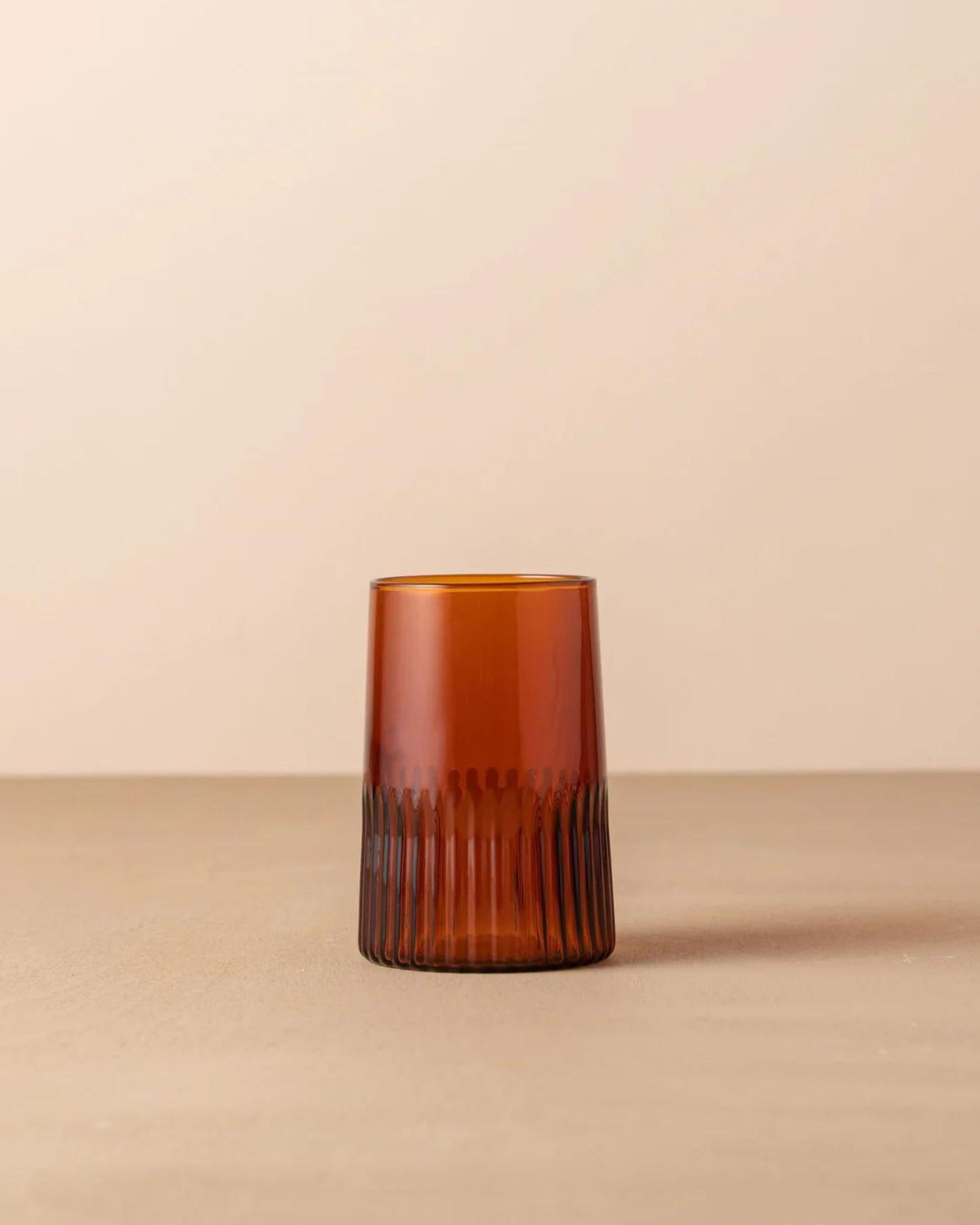 A Saarde KAIROS WATER GLASS, featuring a brown, ribbed design that echoes the elegance of Murano glassware, rests on a neutral surface against a light beige background. This tumbler boasts vertical grooves along its lower half, providing texture and visual interest.